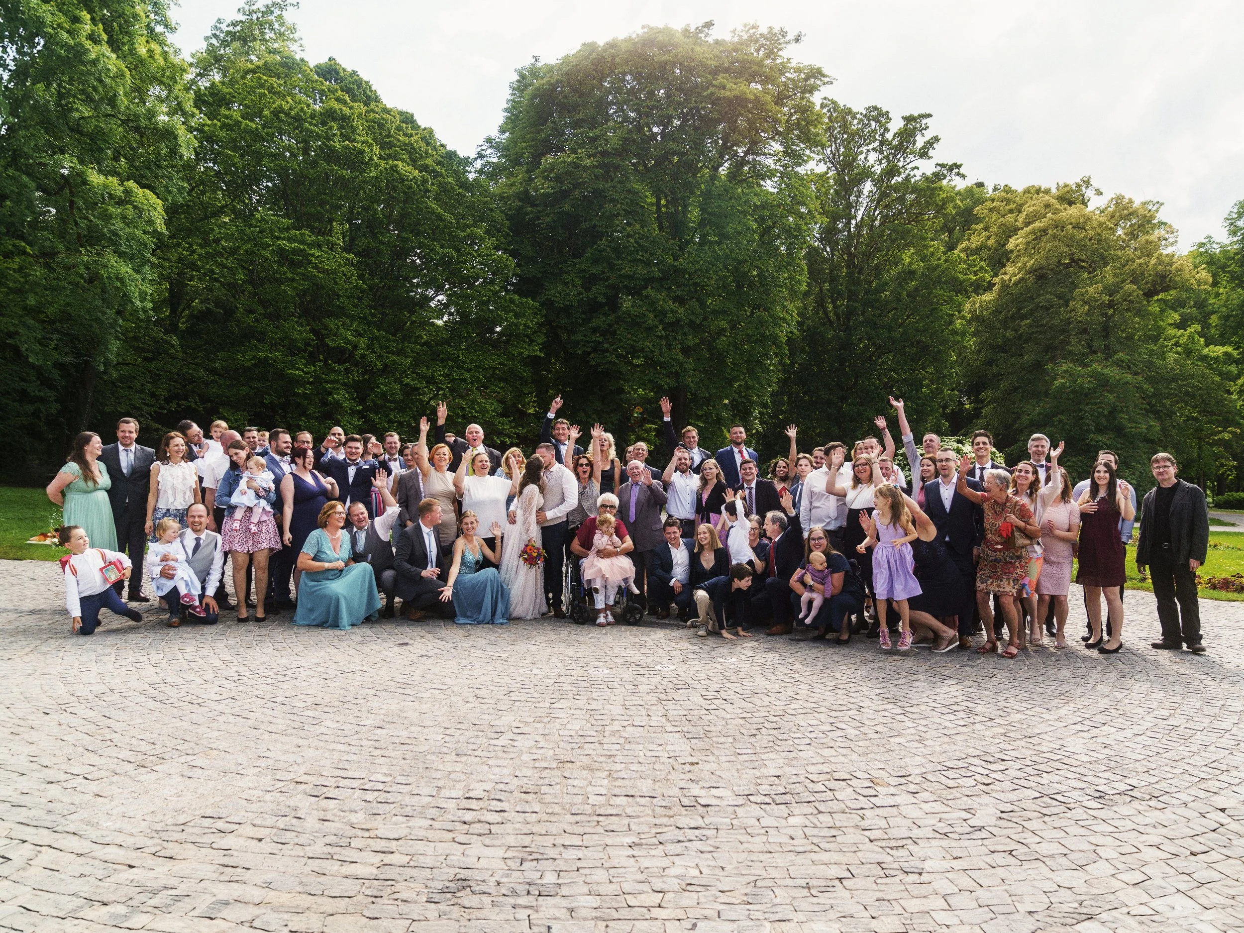 Eine große Gruppe von Menschen, darunter Familien, Freunde und Kinder, steht zusammen im Freien auf einer gepflasterten Fläche, um einen besonderen Anlass zu feiern, mit grünen Bäumen im Hintergrund.