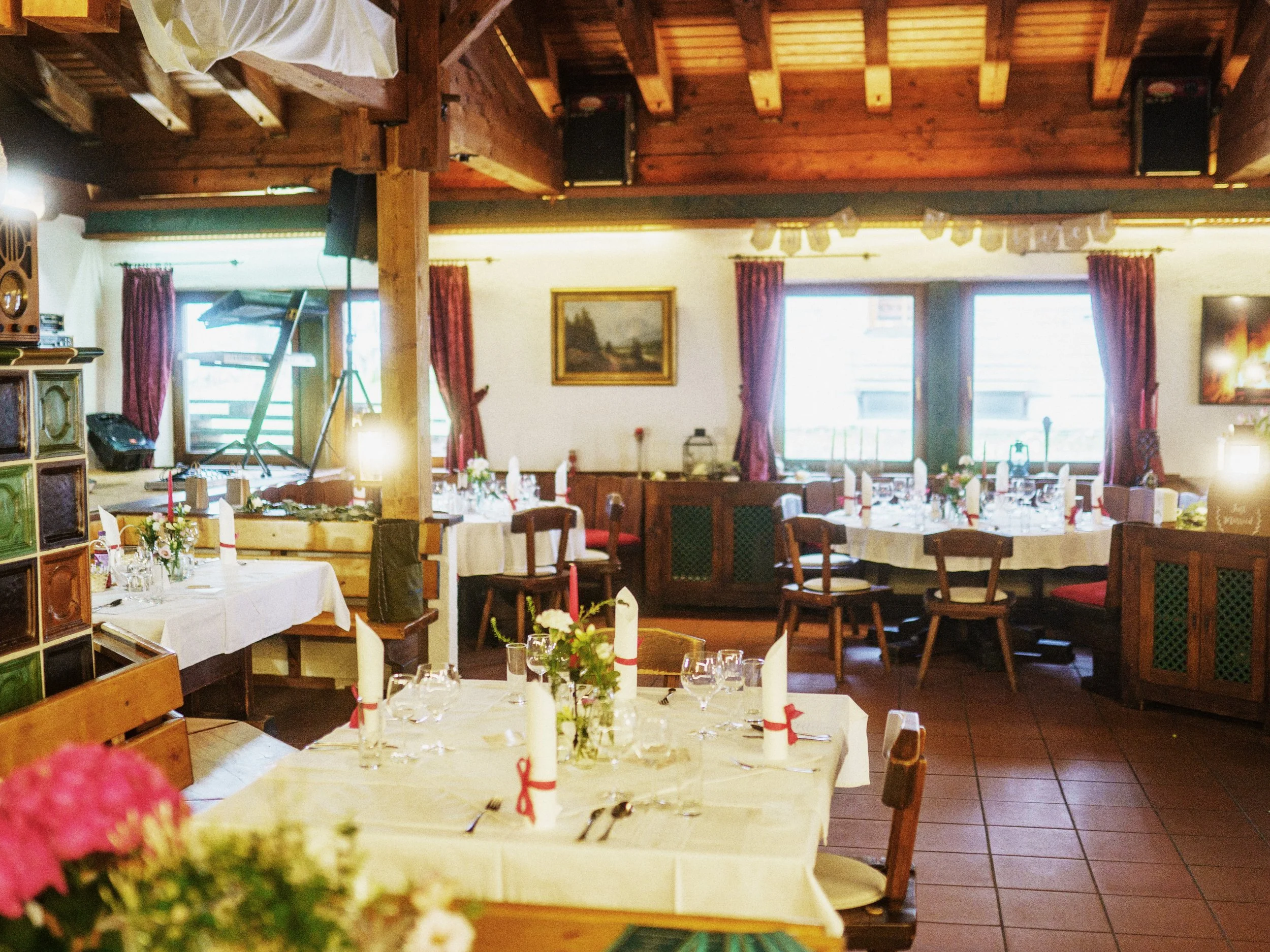 Gemütlich gedeckter Speisesaal in einem rustikalen Restaurant mit Holzdecken, großen Fenstern mit roten Vorhängen und festlich dekorierten Tischen mit Kerzen, Blumen und Besteck.