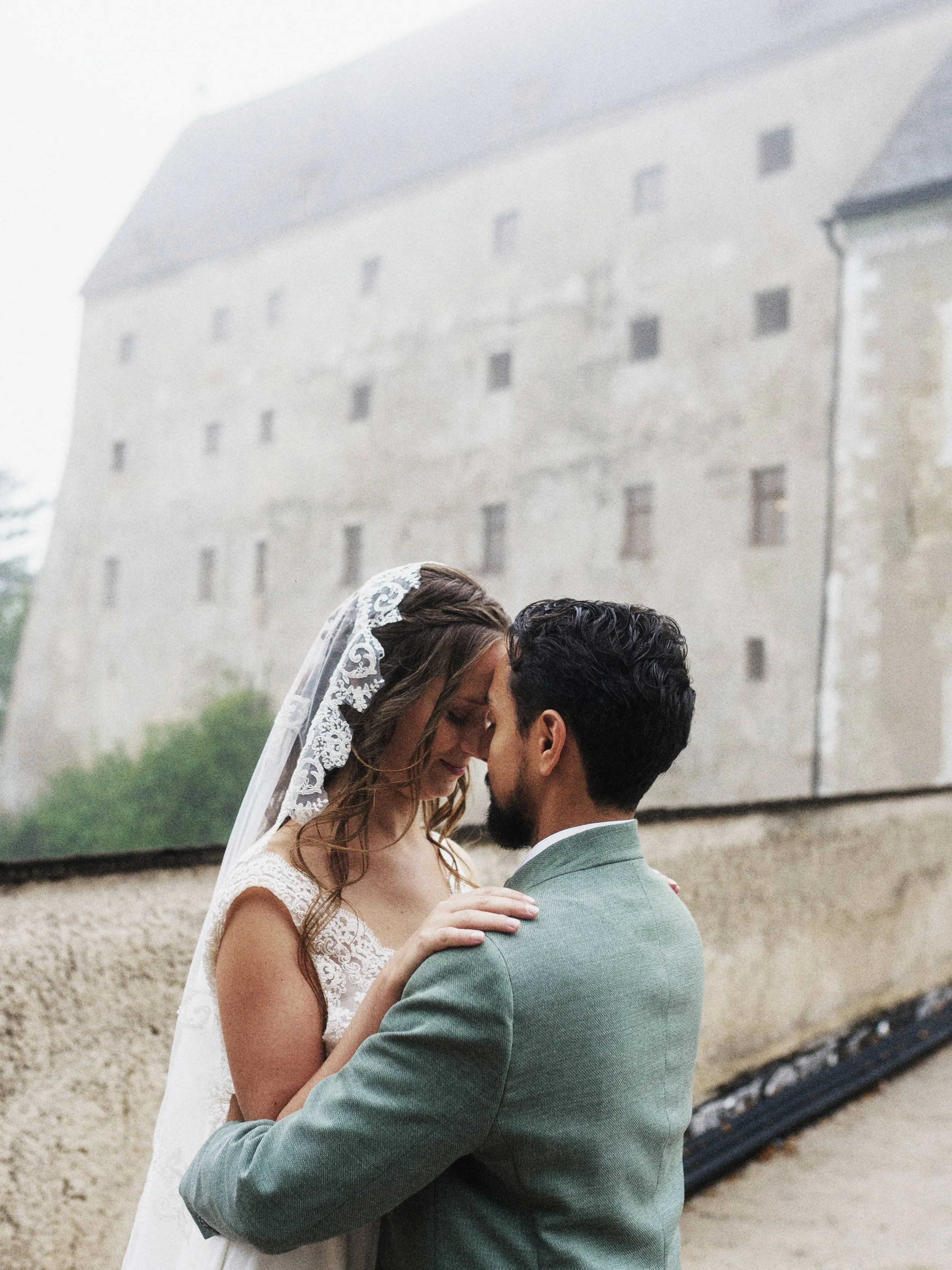 Ein Brautpaar teilt einen intimen Moment vor einer mittelalterlichen Burg, umgeben von natürlicher Umgebung, an einem bewölkten Tag.