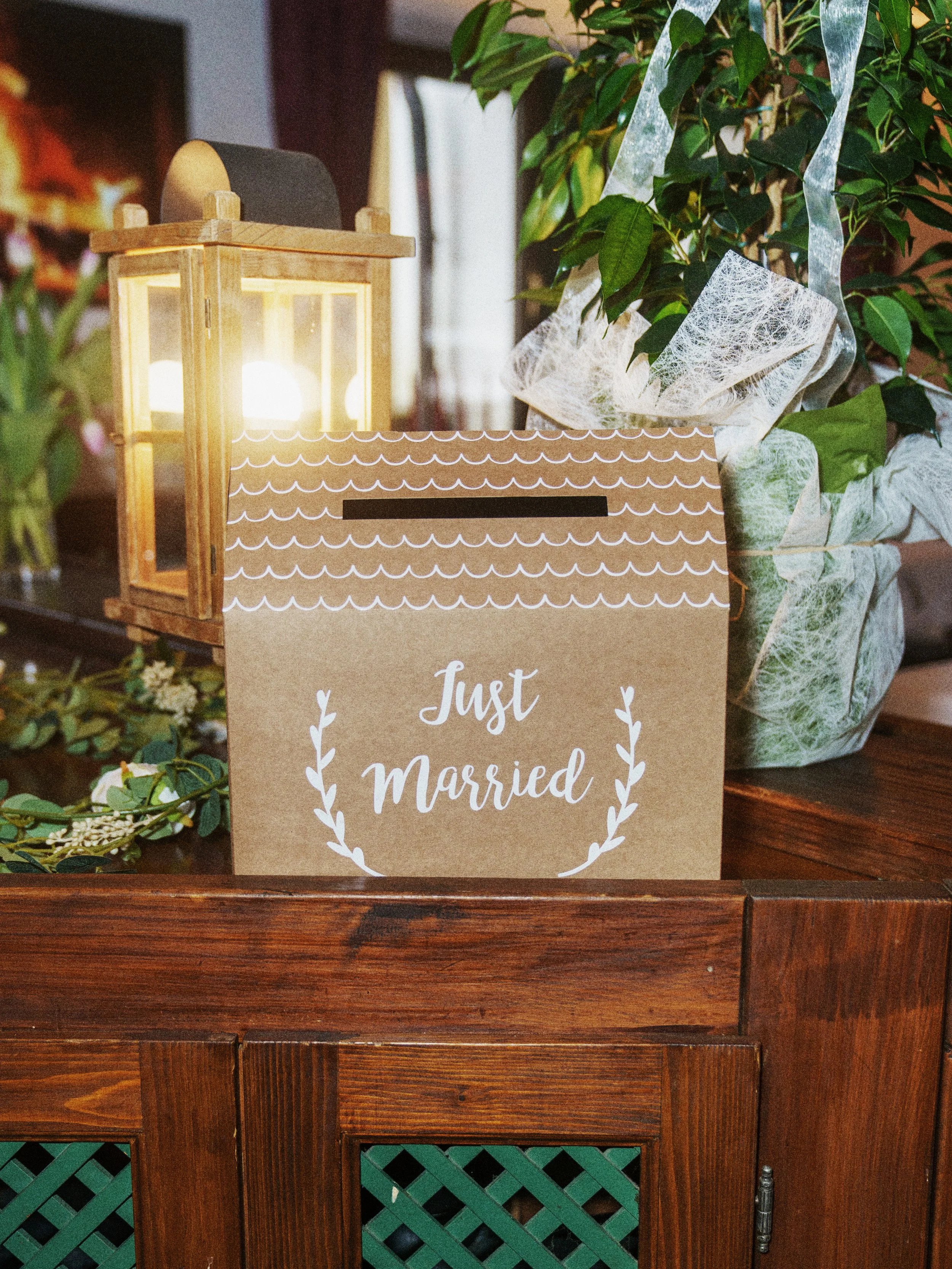 Eine Holztruhe mit einem Schild, auf dem steht 'Just Married', umgeben von Pflanzen und einem Laternenlicht.