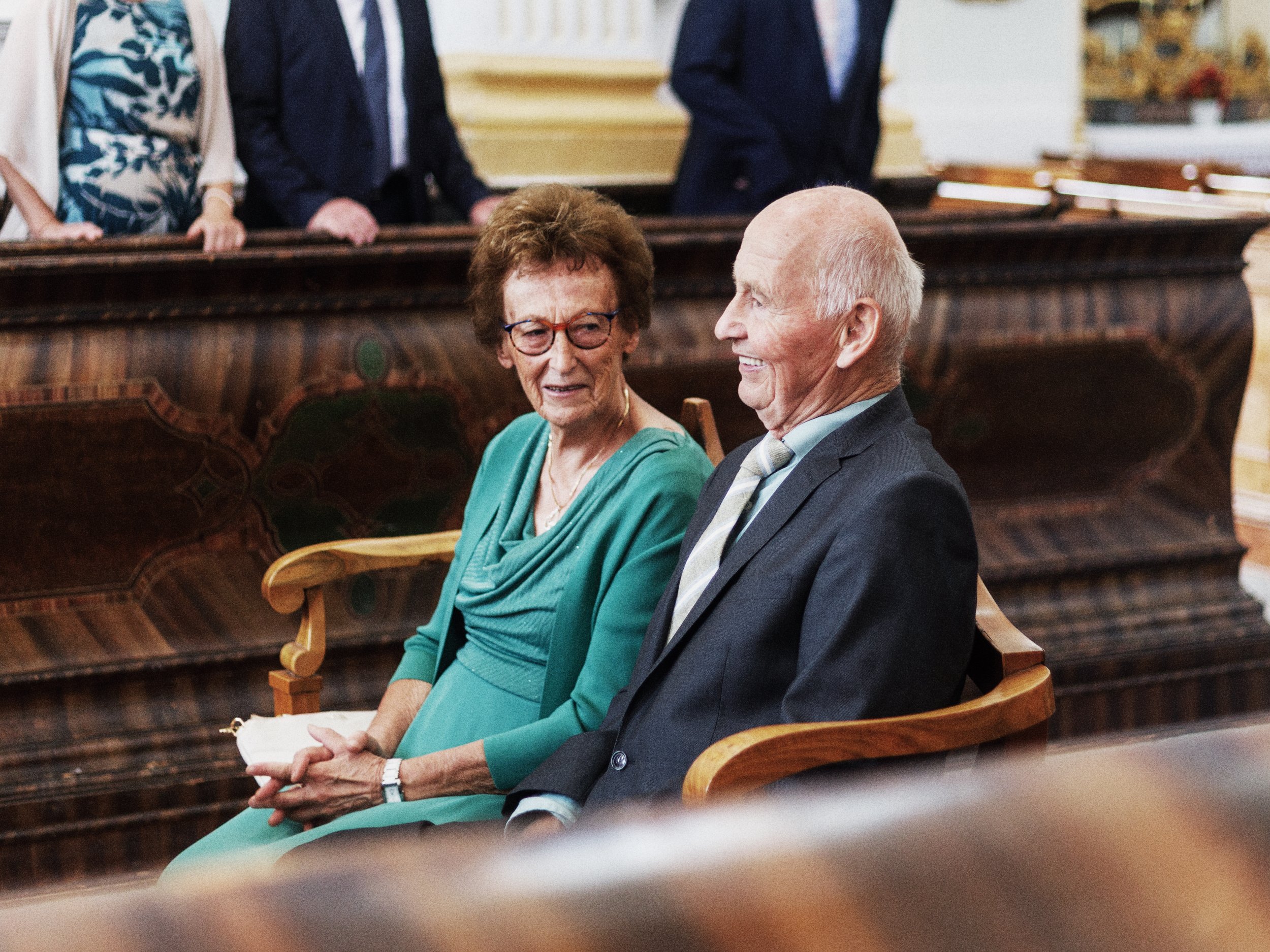 Ein älteres Paar, eine Frau mit Brille in einem grünen Kleid und ein Mann in einem Anzug, sitzen in einem Raum und lachen miteinander bei einer Feier oder Zeremonie.