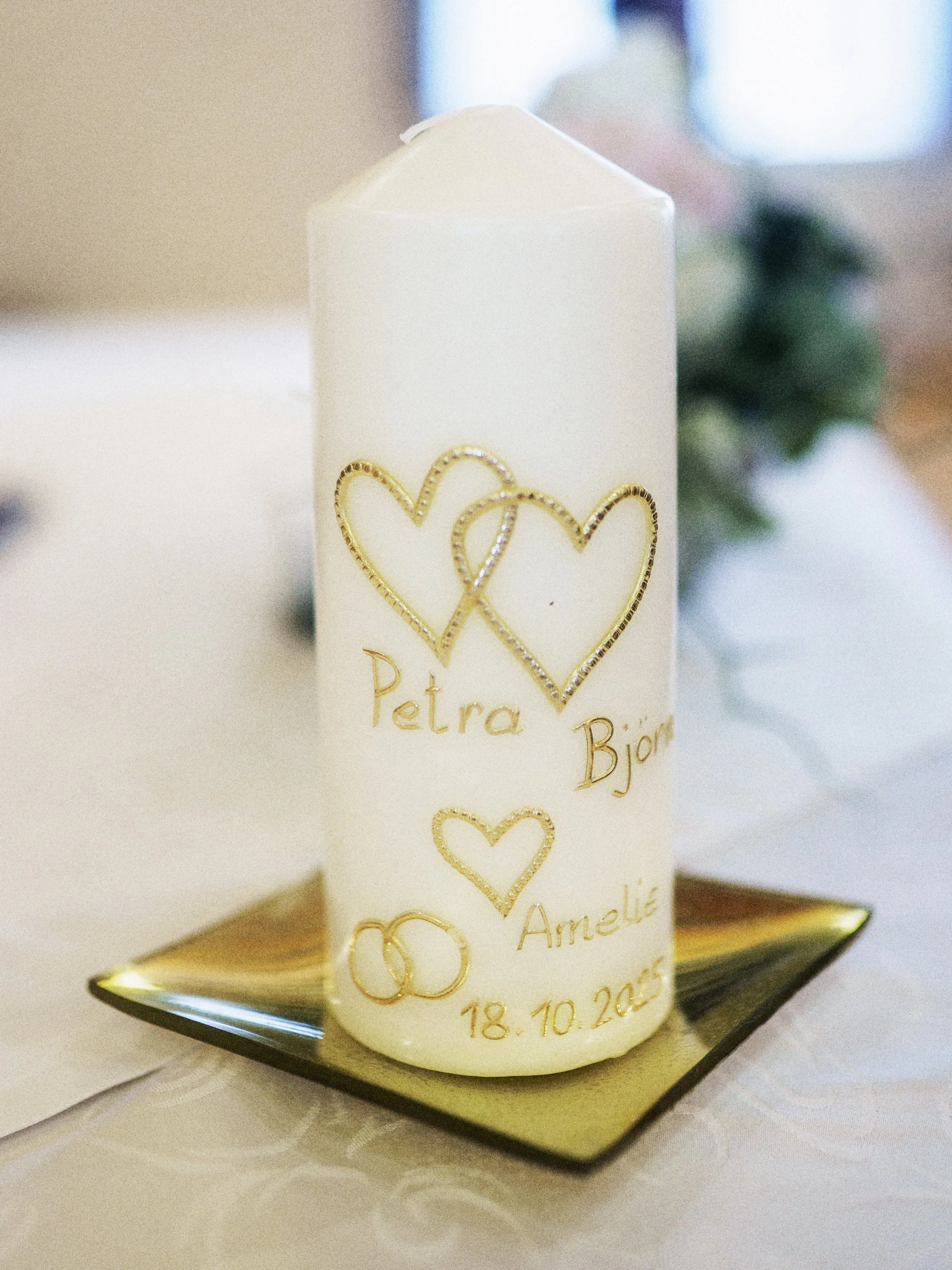 Eine weiße Kerze mit goldenen Herzen, Namen und Hochzeitsdatum darauf, auf einem goldenen Teller, im Hintergrund unscharf.