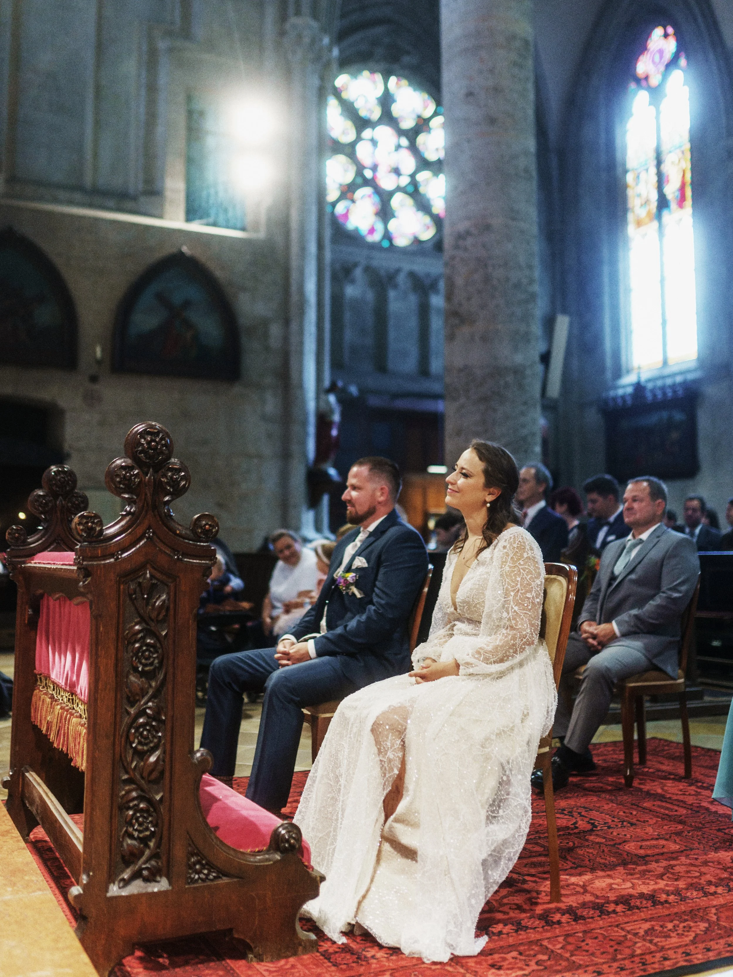 Ein Hochzeitspaar sitzt während einer Zeremonie in einer Kirche, umgeben von Gästen, mit bunten Kirchenfenstern im Hintergrund.