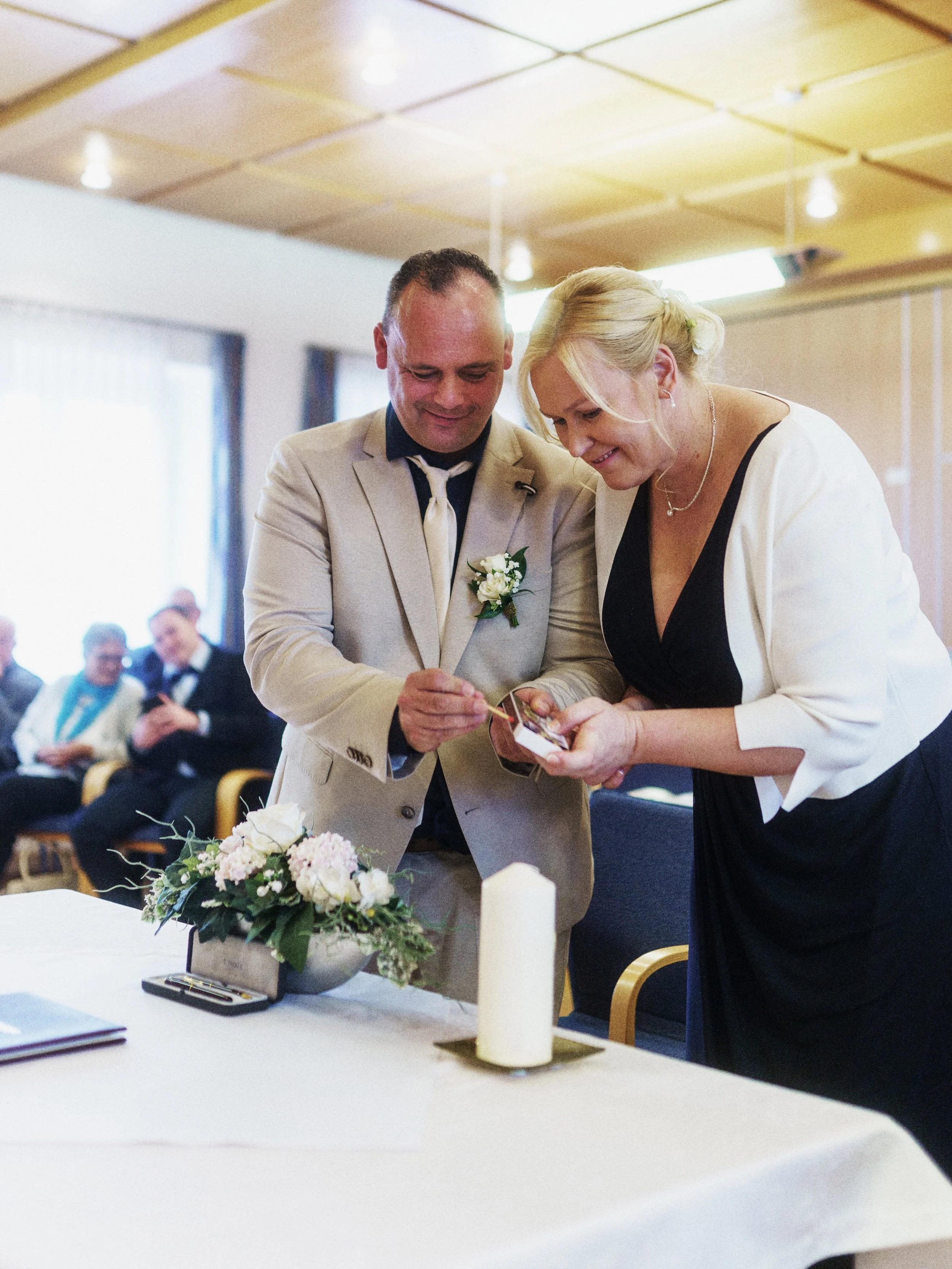 Ein Hochzeitspaar bei der Zeremonie, die gemeinsam einen Ring tauschen, im Vordergrund ein Tisch mit Blumen und einer Kerze, im Hintergrund Gäste, die zuschauen.