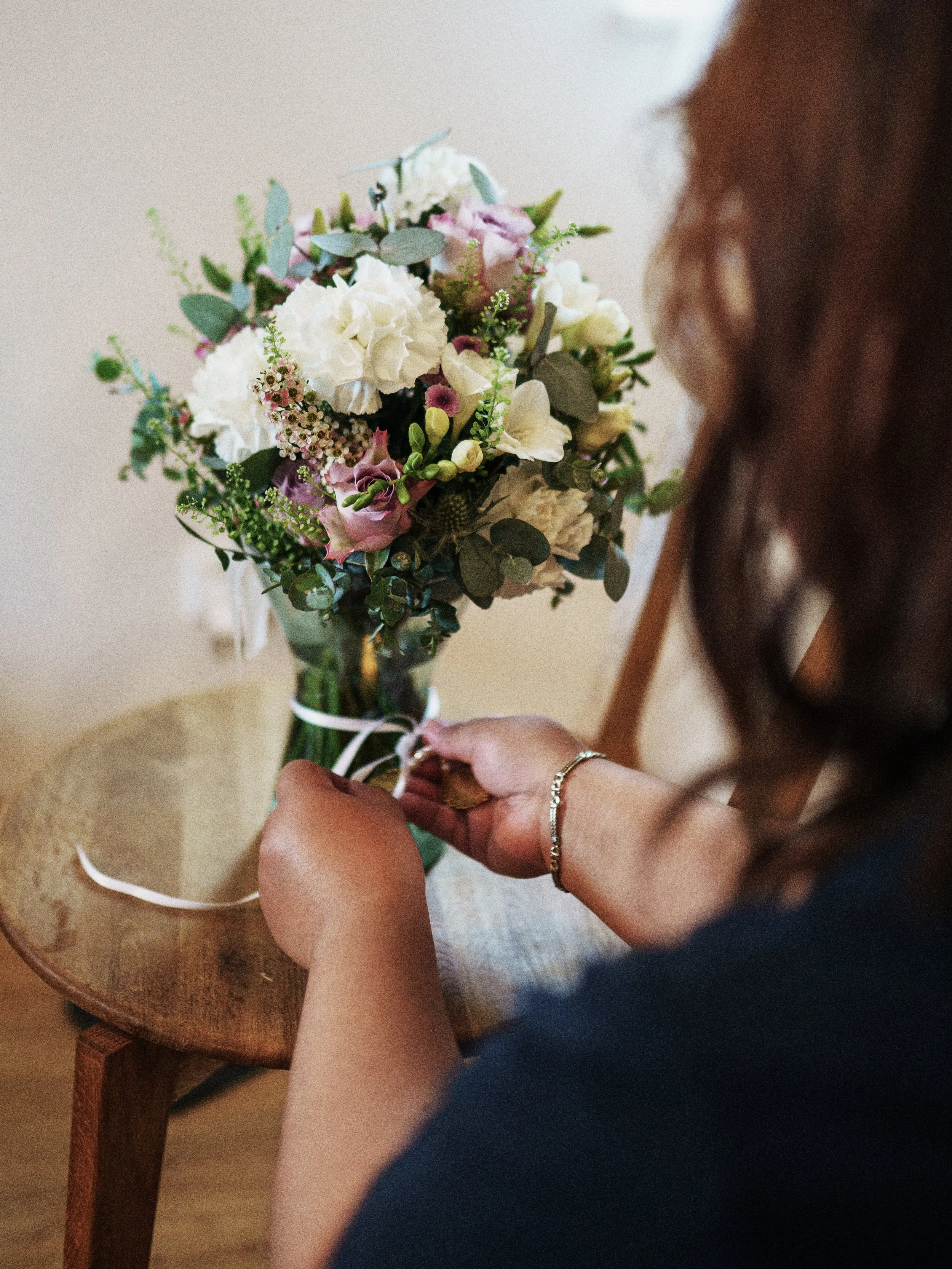 Eine Person bindet einen Blumenstrauß auf einem kleinen Holzstuhl. Der Blumenstrauß enthält weiße, rosa und grüne Blumen und ist mit einer weißen Schleife verziert.