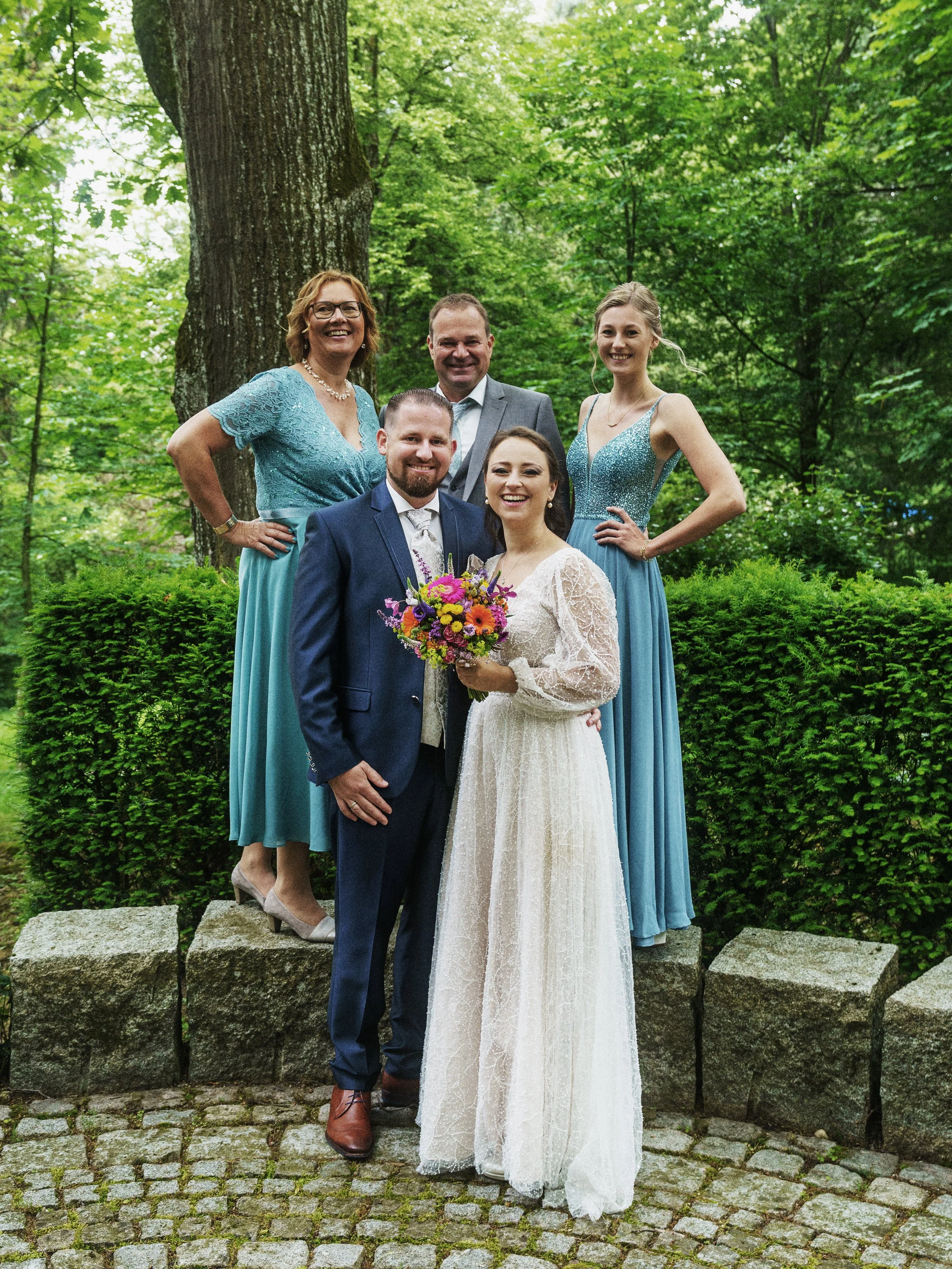 Hochzeitspaar mit drei Gästen in einem grünen Park, lachend, mit Blumen, vor großen Bäumen und einer Hecke.