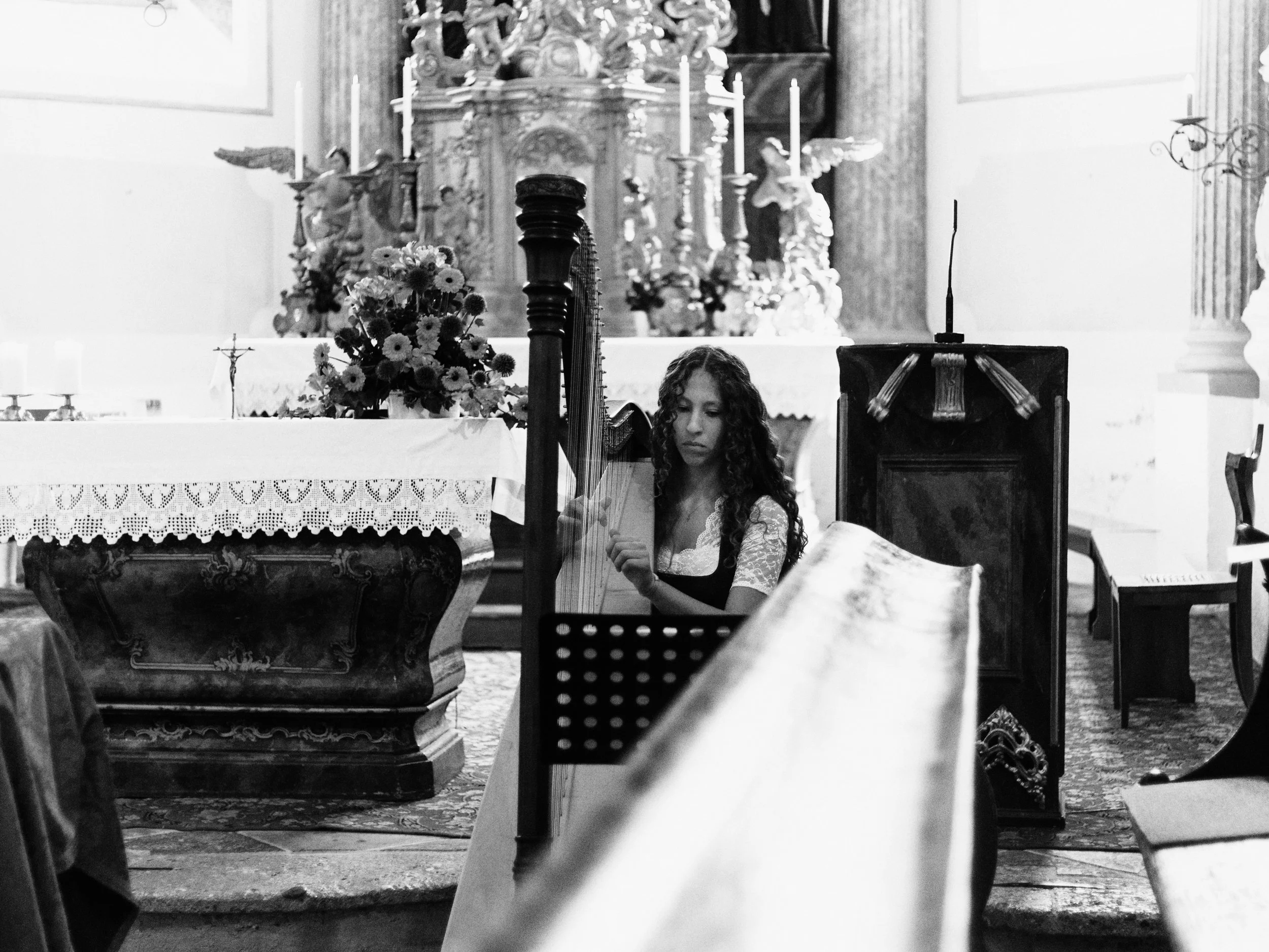Eine junge Frau mit langen, lockigen Haaren spielt Harfe im Kirchenschiff bei einer Zeremonie, im Hintergrund Altar mit Kerzen, Blumen und religiösen Figuren, alles in schwarz-weiß.