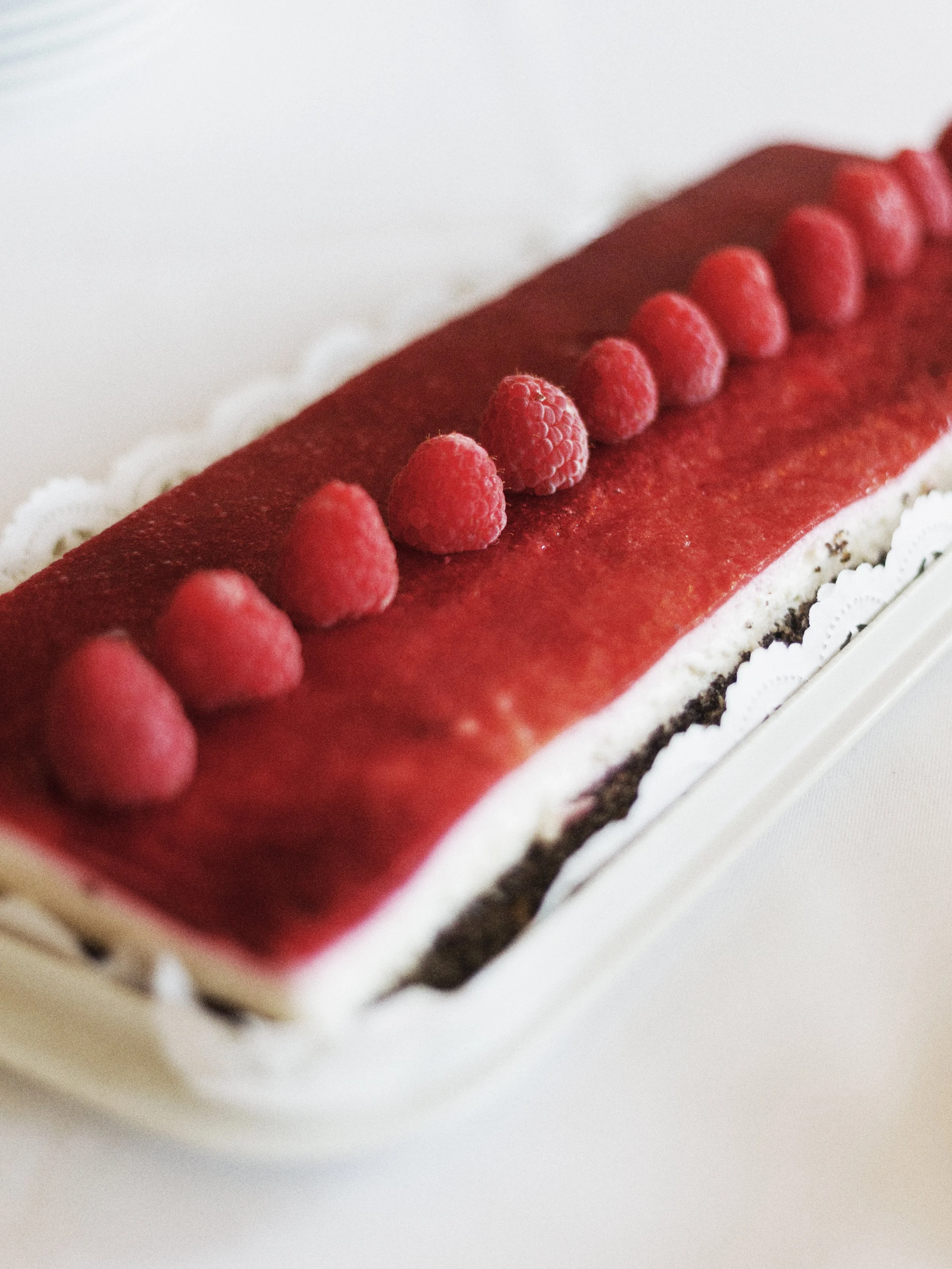 Ein rechteckiger Erdbeerkuchen mit Himbeeren auf der Oberseite, auf einem weißen Untersetzer.