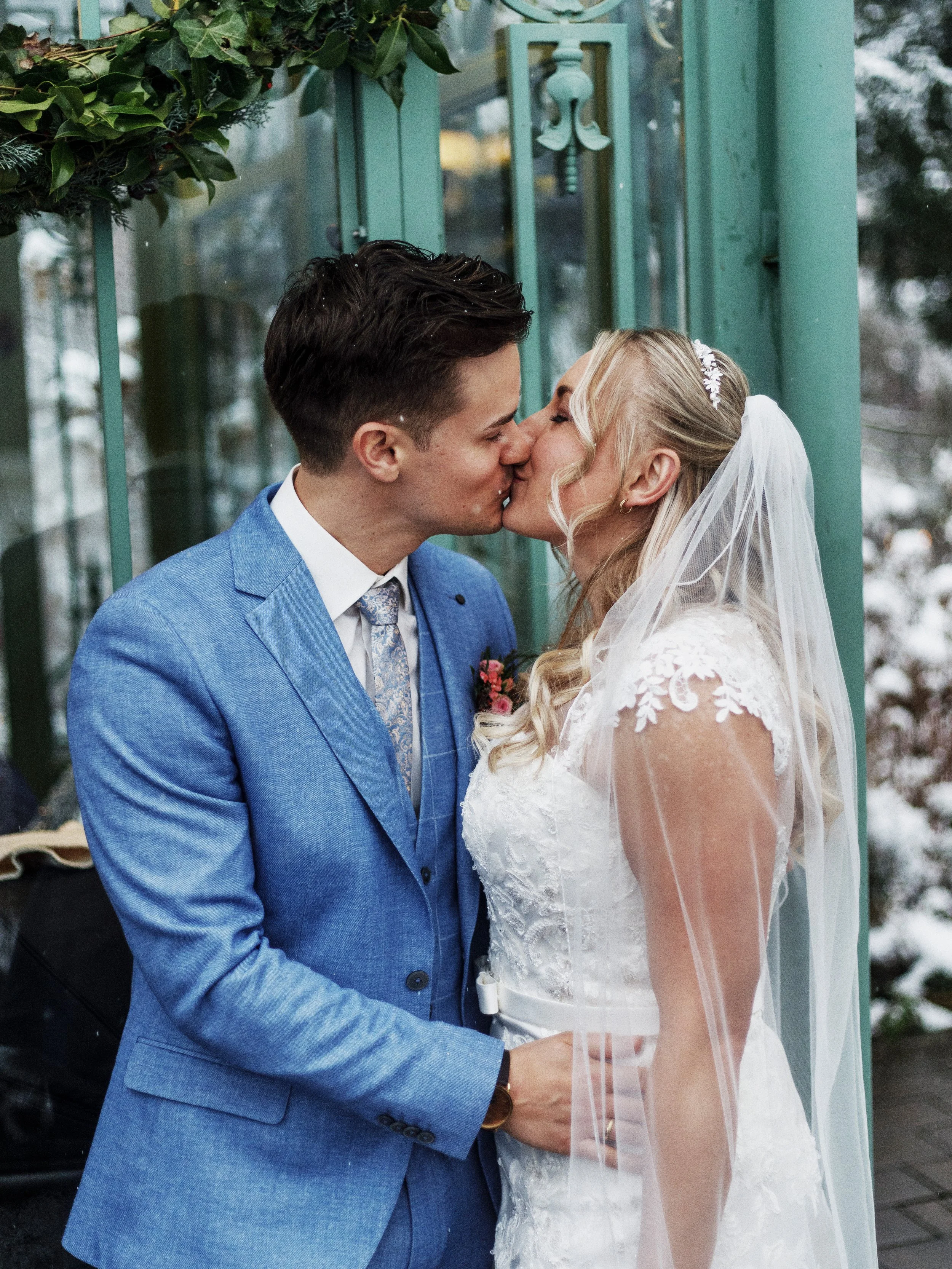 Verliebtes Brautpaar küsst sich vor einem grünen Türrahmen, sie trägt ein weißes Hochzeitskleid mit Spitzendetails und Tüll, er trägt einen blauen Anzug mit Krawatte, im Hintergrund ist Schnee sichtbar.