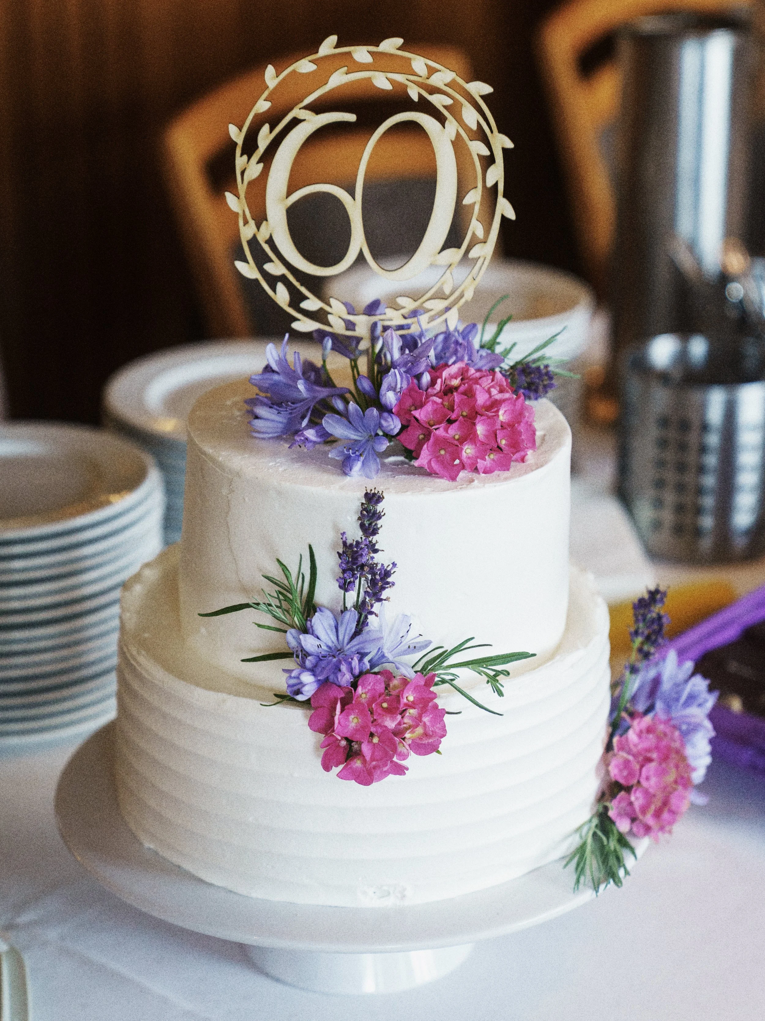 Hochzeitstorte mit weißen Schichten, dekoriert mit pinken und lila Blumen, mit einem goldenen Tortenstecker mit der Zahl 60.