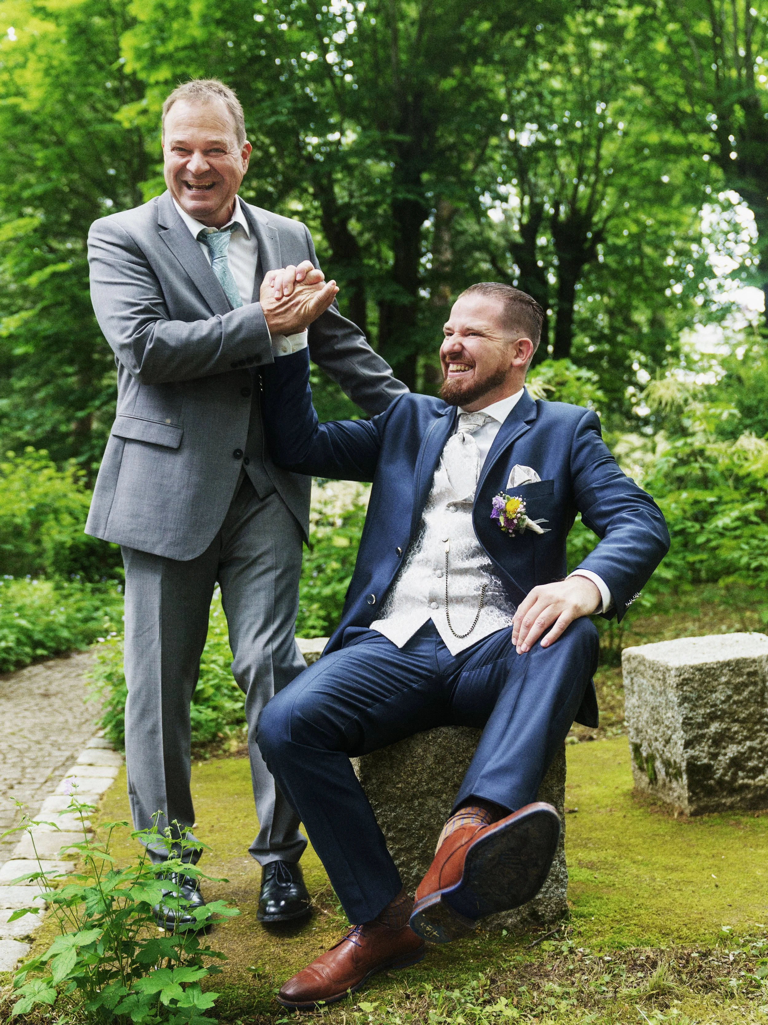 Zwei Männer in Anzügen, einer sitzt auf einem Stein und der andere steht neben ihm, beide lachen und halten sich an der Hand, im Hintergrund ein grüner Park.