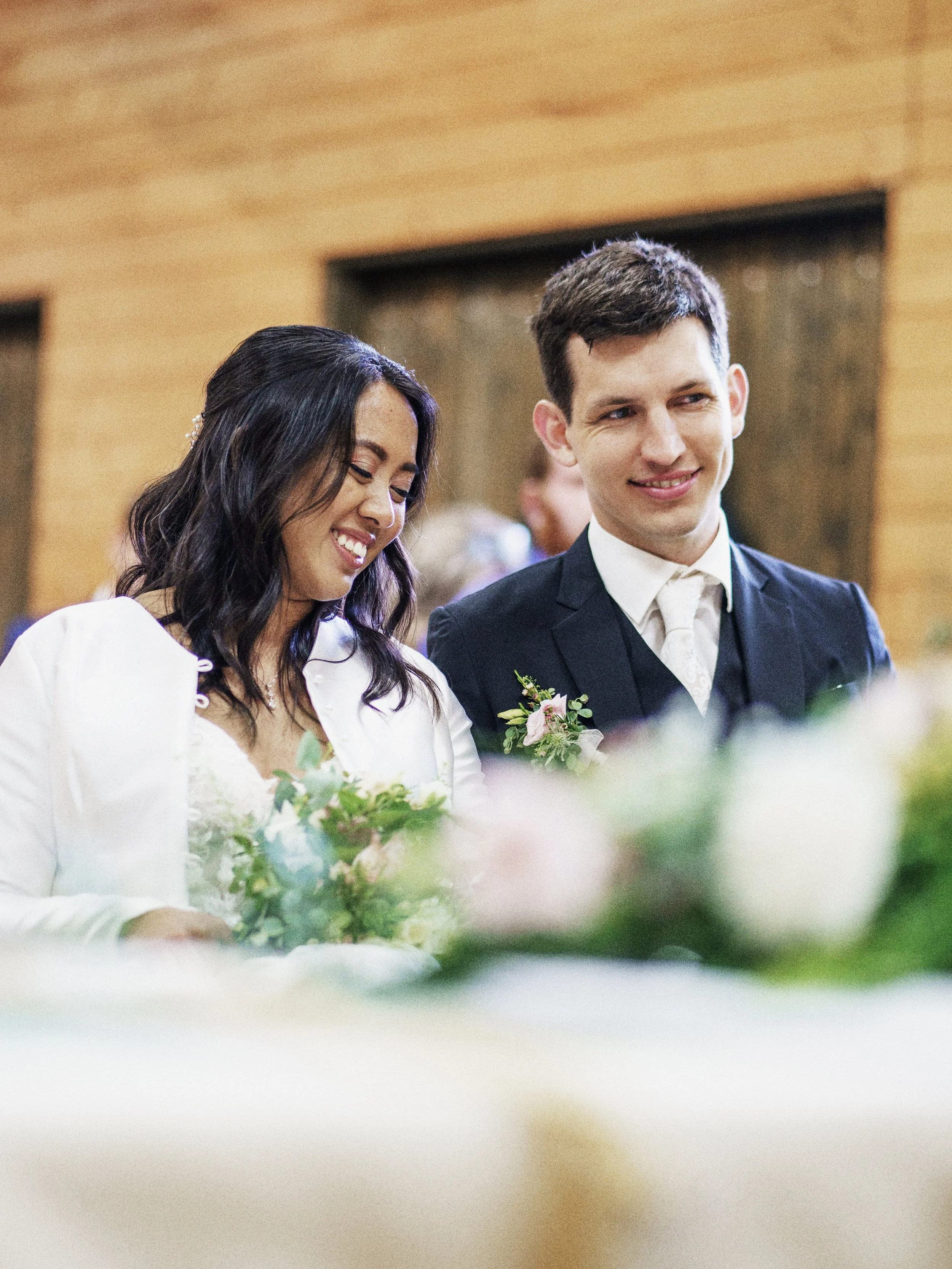 Ein Brautpaar bei der Hochzeit, lachend und interessiert an den Blumen auf dem Tisch.