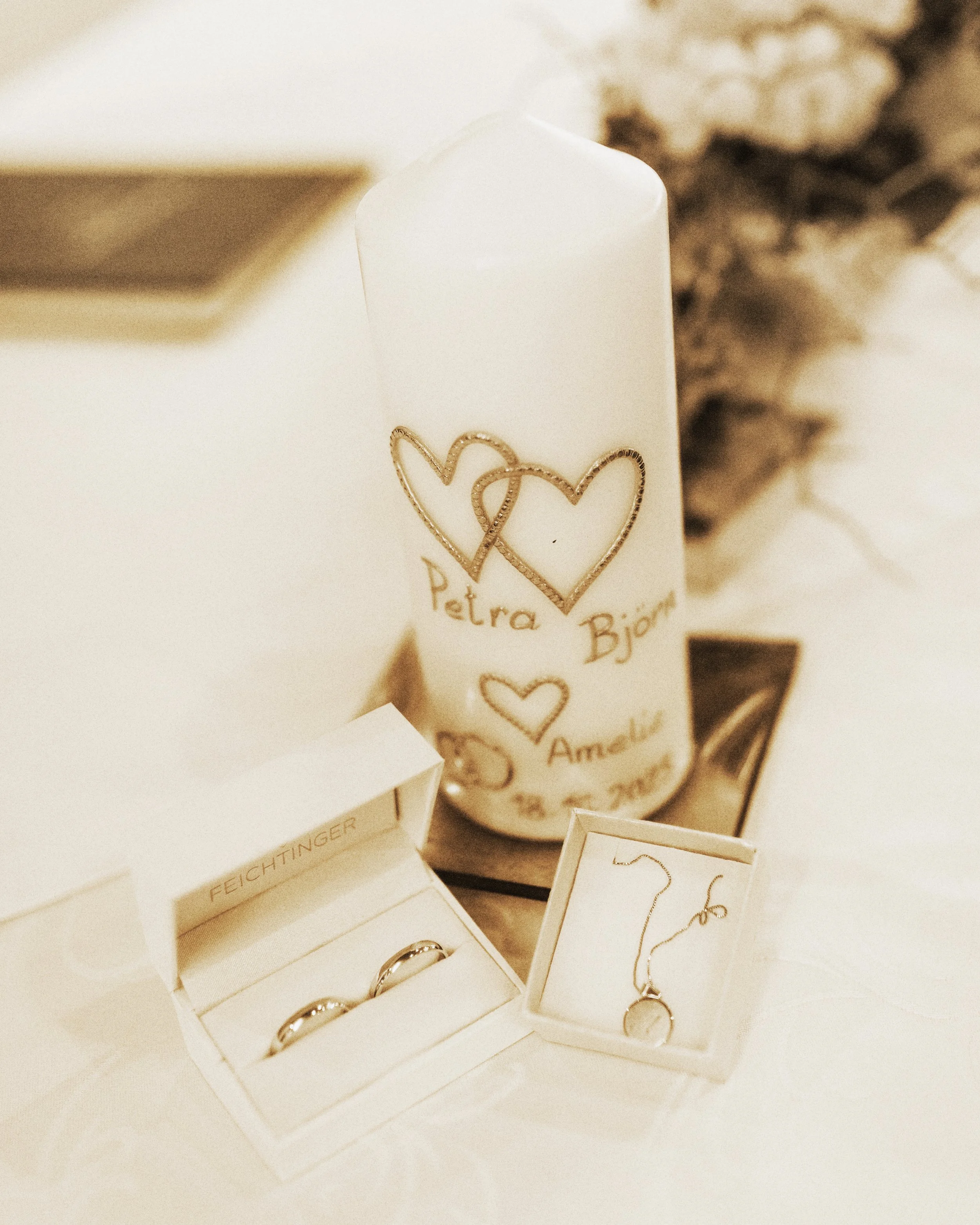 Hochzeitskerze mit goldenen Herzen und Namen Petra und Björn, daneben zwei Eheringe in einer weißen Box und eine Halskette in einer kleinen Schachtel.