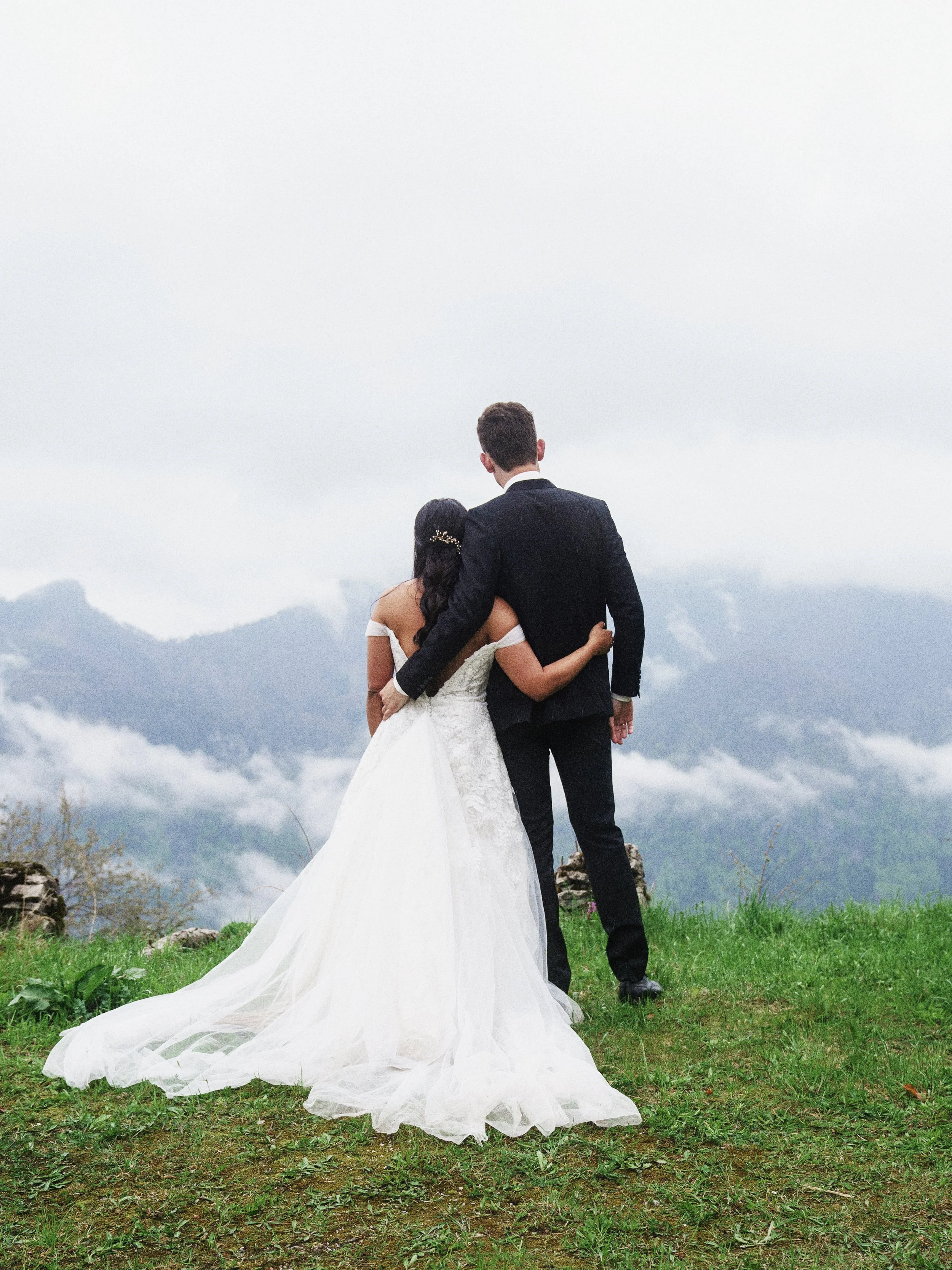 Braut und Bräutigam stehen auf grünem Rasen, umarmt vor Bergen und Wolken, Blick nach hinten.