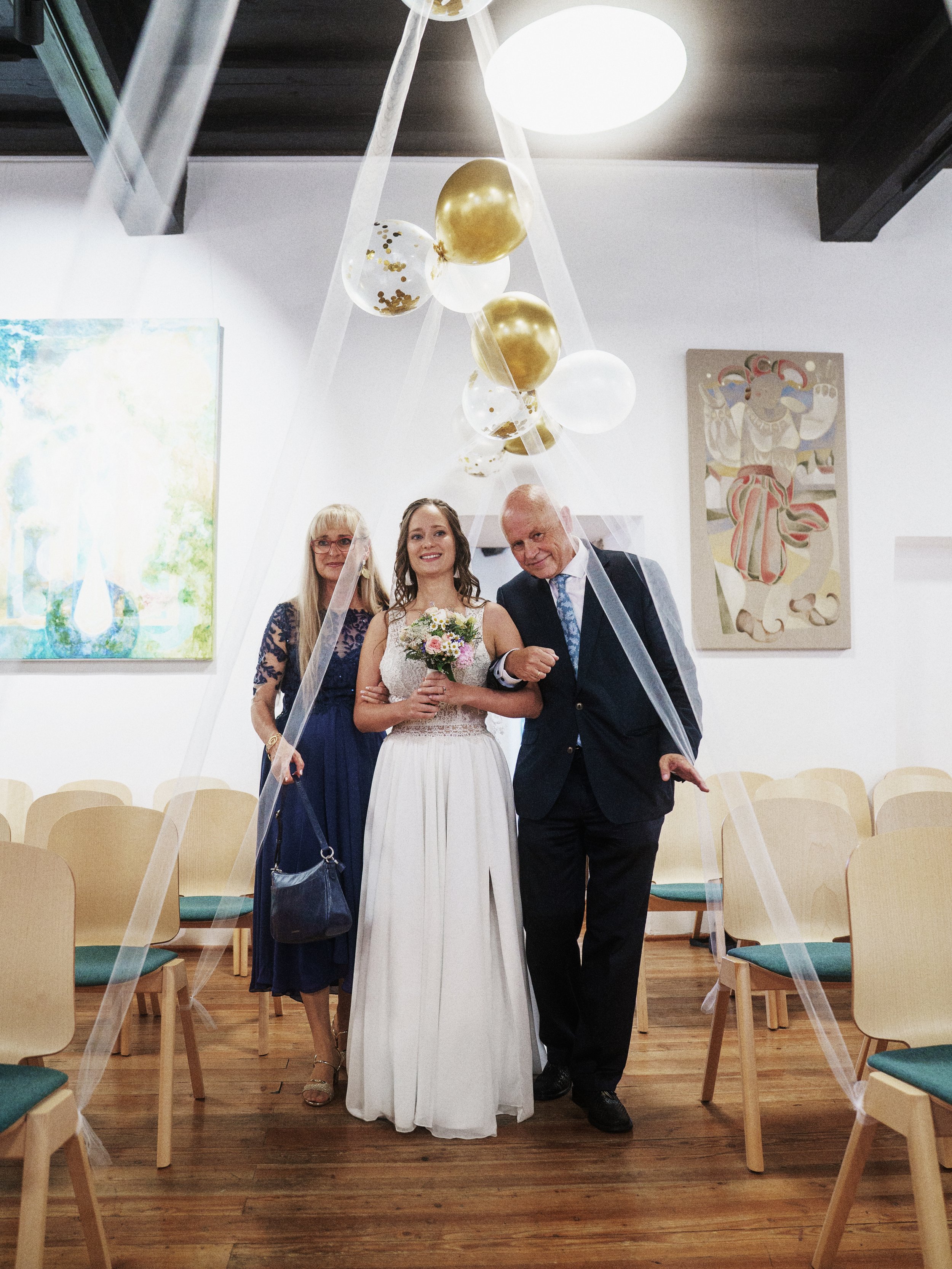 Ein junges Brautpaar wird von den Eltern, in einem Raum mit moderner Kunst an den Wänden begleitet. Es handelt sich um eine Hochzeitsfeier.