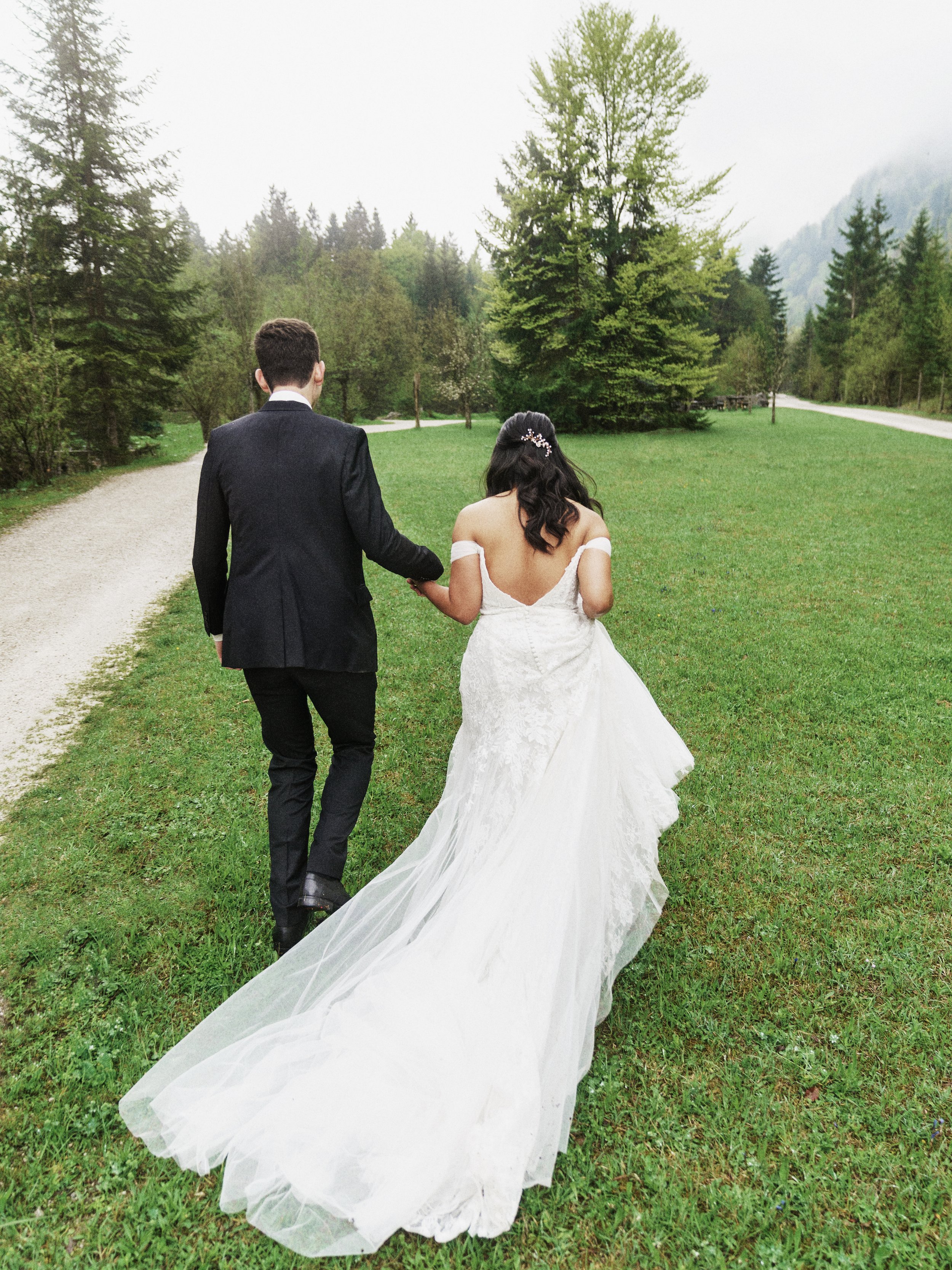 Ein Brautpaar, das zusammen durch eine grüne Wiesenlandschaft in den Bergen spaziert, mit bewölktem Himmel im Hintergrund.