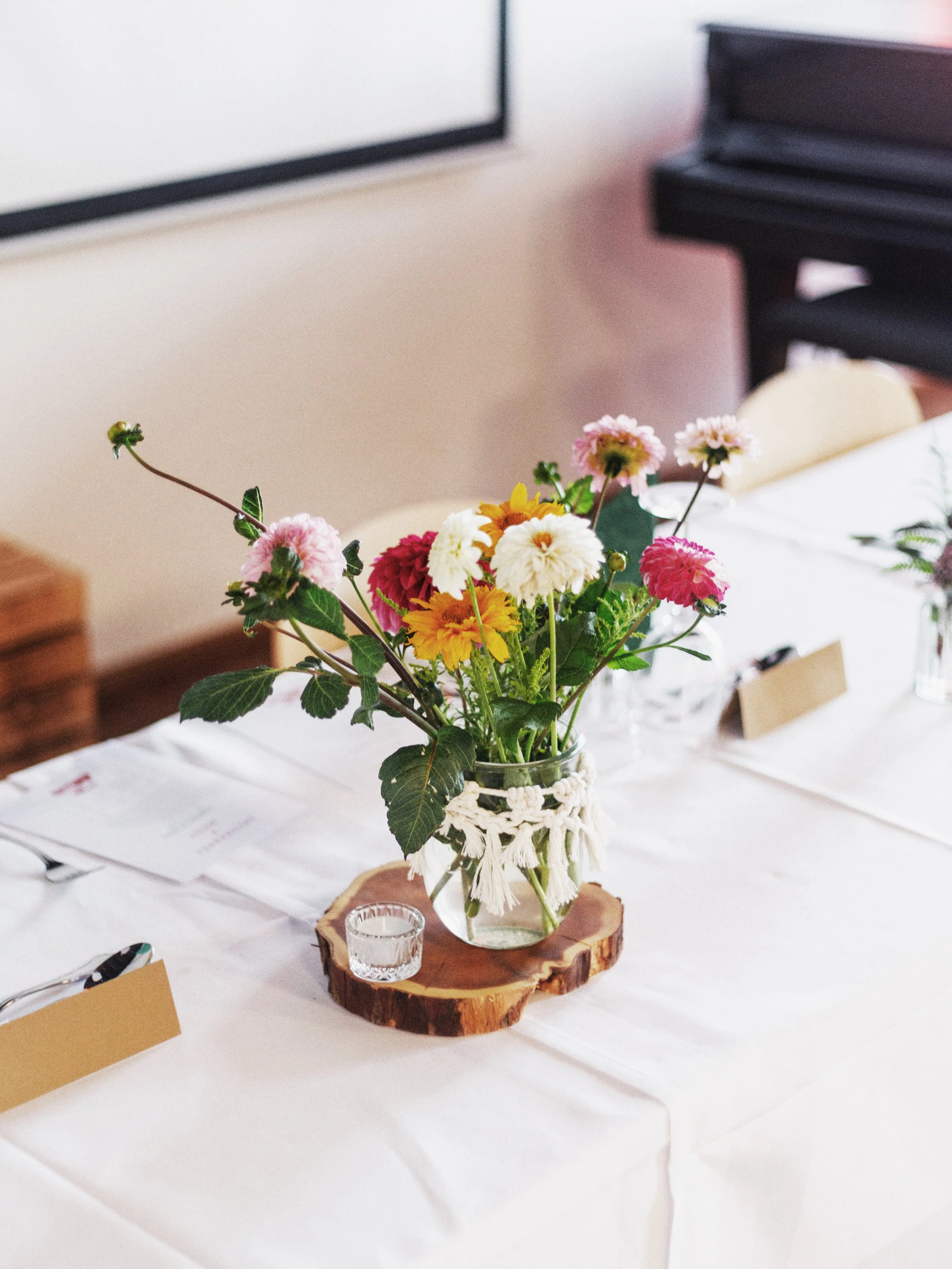 Blumenstrauß in einem Glas mit Makramee-Deckel auf einem Holzscheit auf einem weißen Tisch bei einer Veranstaltung.
