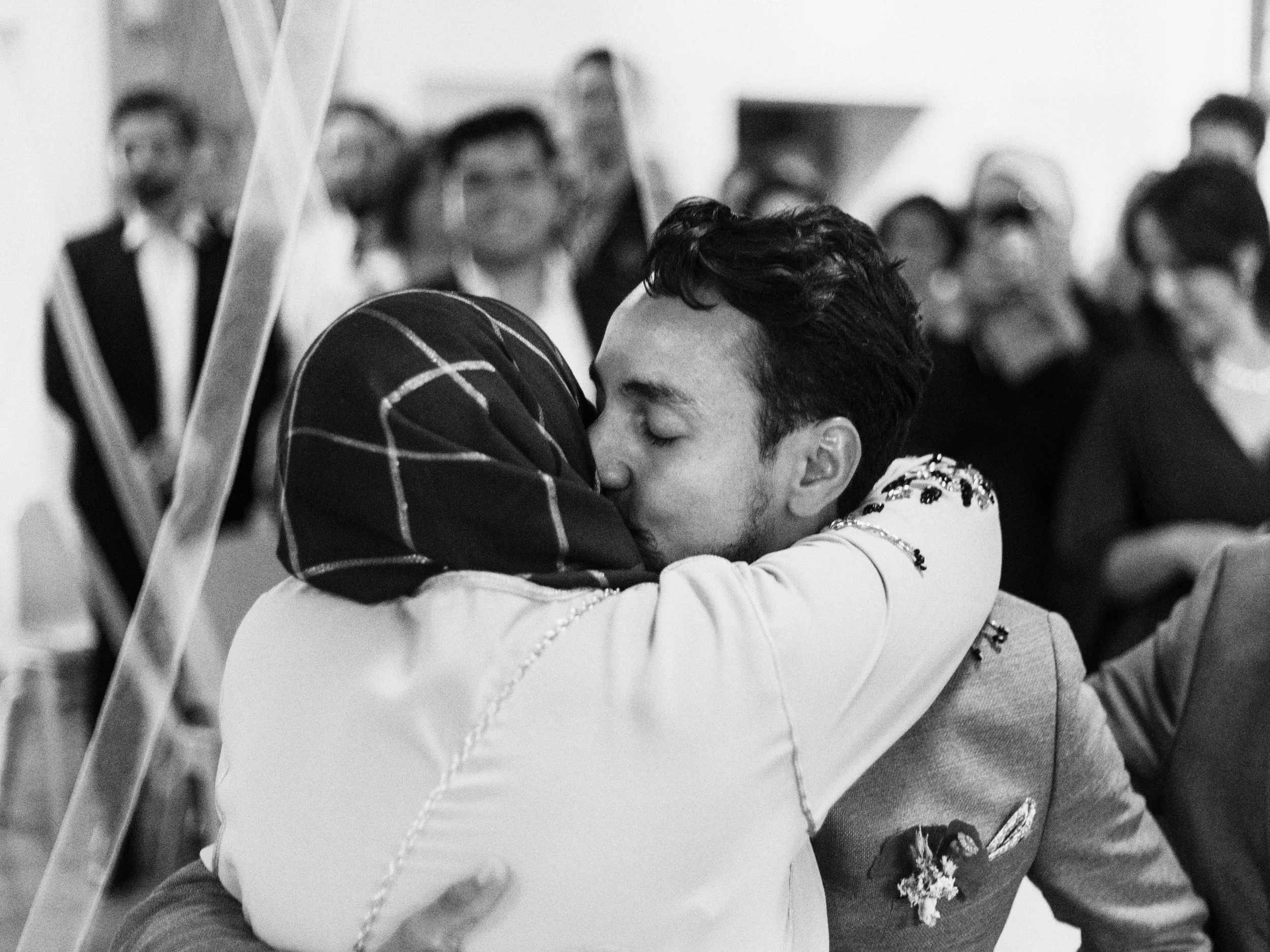 Ein Bräutigam und seine Mutter umarmen sich beim Fest, im Hintergrund stehen weitere Gäste, das Bild ist schwarz-weiß.