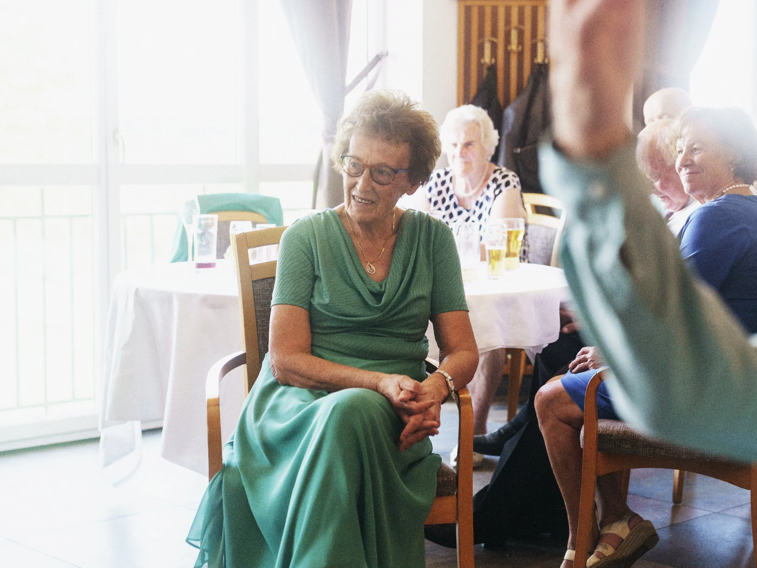 Eine ältere Frau in einem grünen Kleid sitzt an einem Tisch bei einer Feier, umgeben von weiteren Gästen, im Hintergrund ein Fenster mit Tageslicht.
