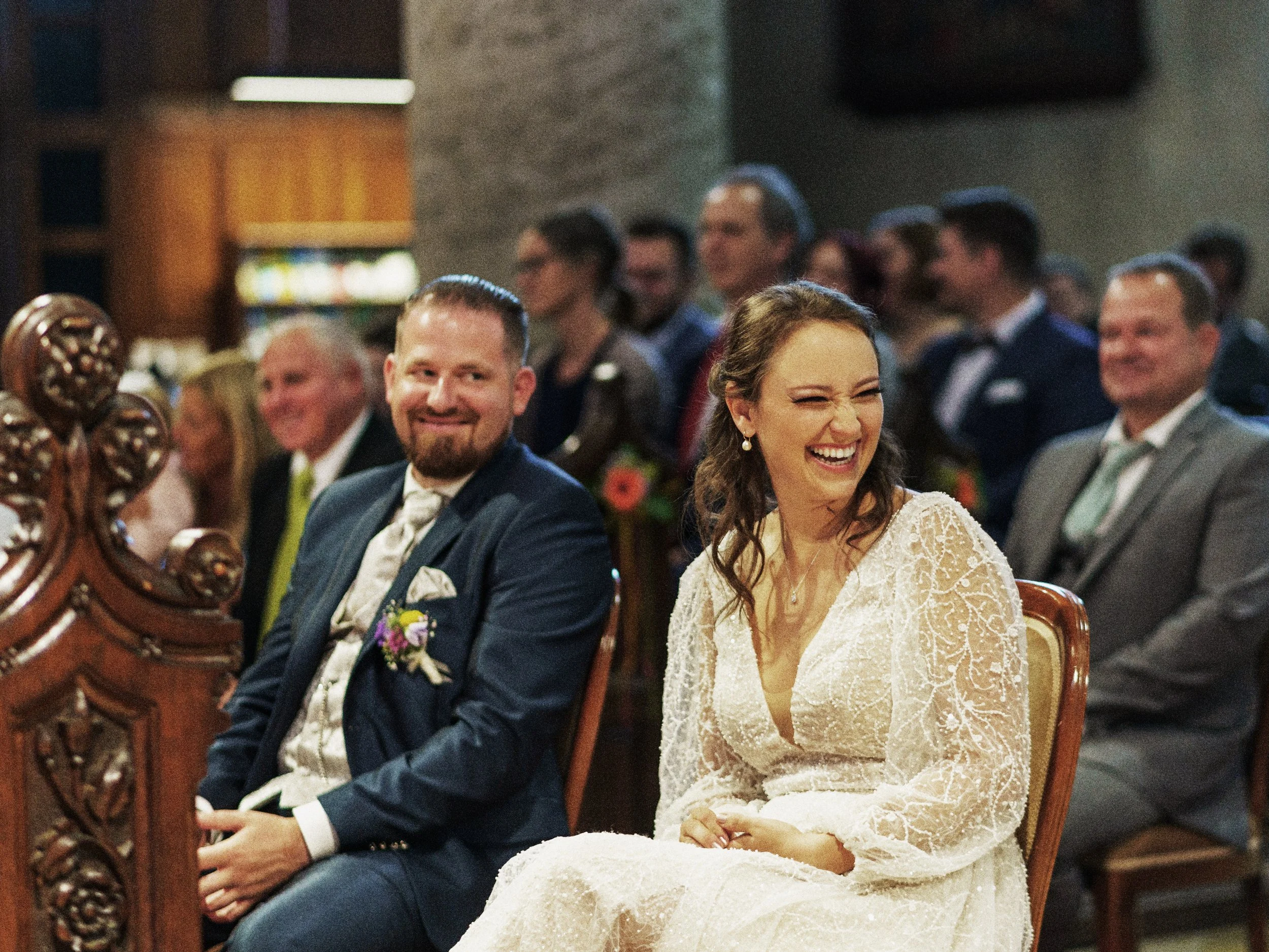 Lächelnder Braut und Bräutigam sitzen bei ihrer Hochzeit, umgeben von Gästen in einem festlichen Raum.