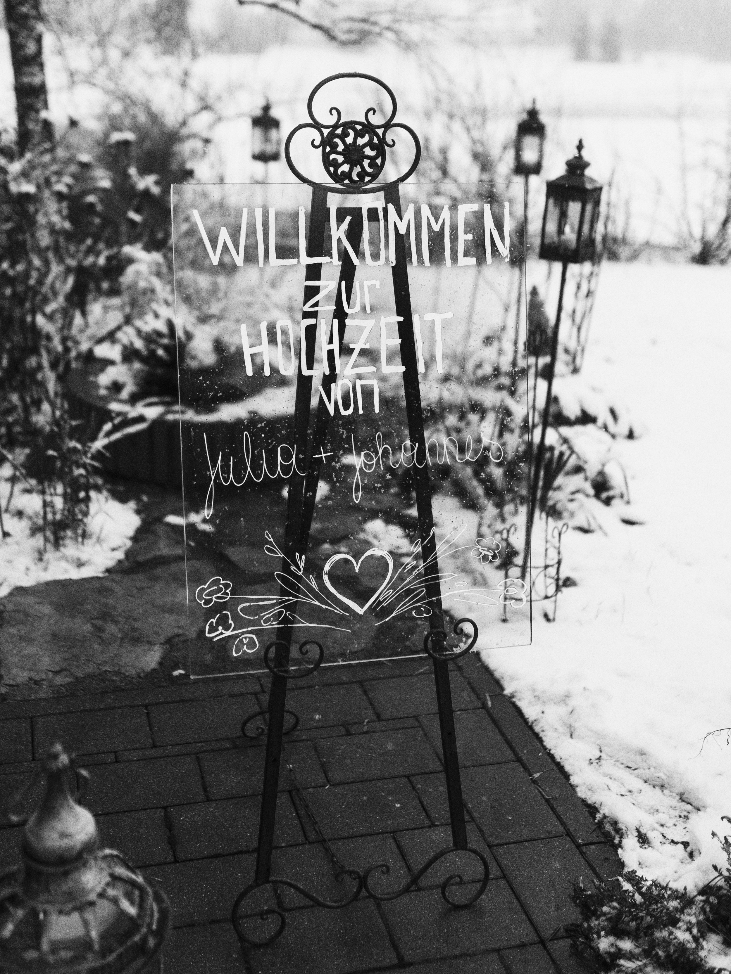 Ein Willkommensschild auf einem Ständer im Schnee, mit handgeschriebenem Text auf Glas, das vor einem Garten mit Laternen steht.