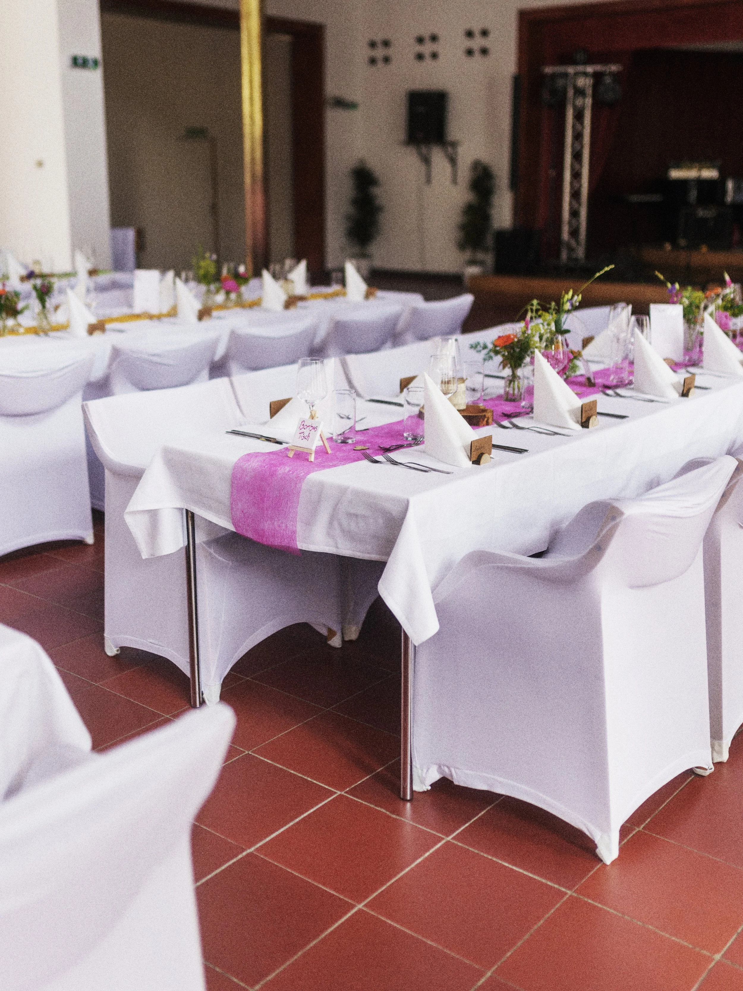 Tisch bei Feier oder Veranstaltung mit weißen Tischdecken, Kerzen, Weingläsern, Besteck, weißen Servietten und bunten Blumen als Dekoration, in einem Veranstaltungsraum mit Bühne im Hintergrund.