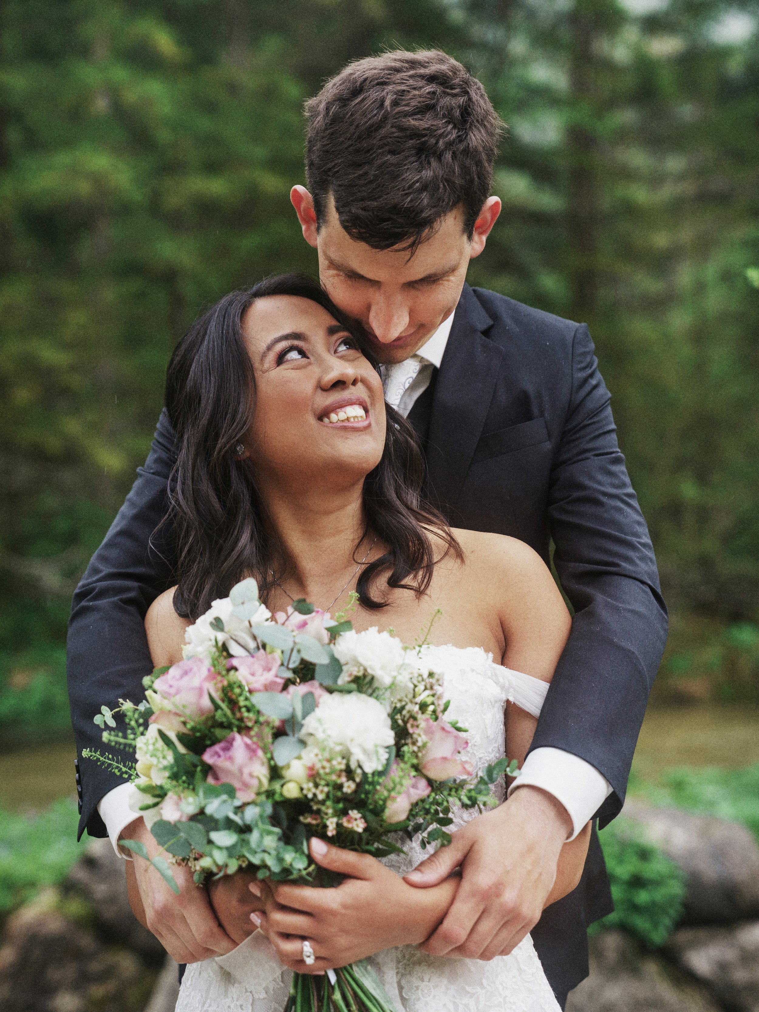 Ein glückliches Brautpaar umarmt sich in einem Wald, die Braut hält einen Blumenstrauß. Der Mann trägt einen Anzug, die Frau ein weißes Hochzeitskleid.