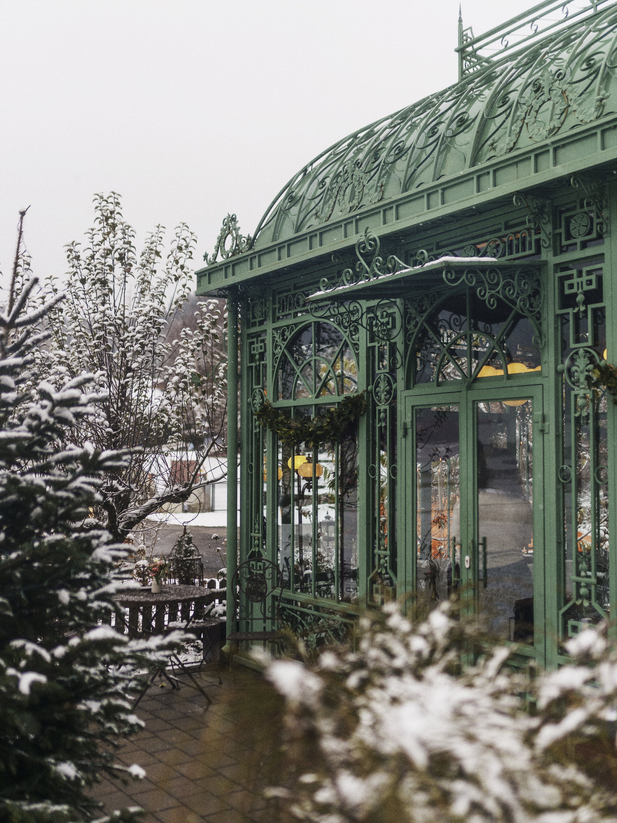 Grünes viktorianisches Gartenhäuschen mit verzierter Metallkonstruktion im Winter, umgeben von schneebedeckten Bäumen und Pflanzen.