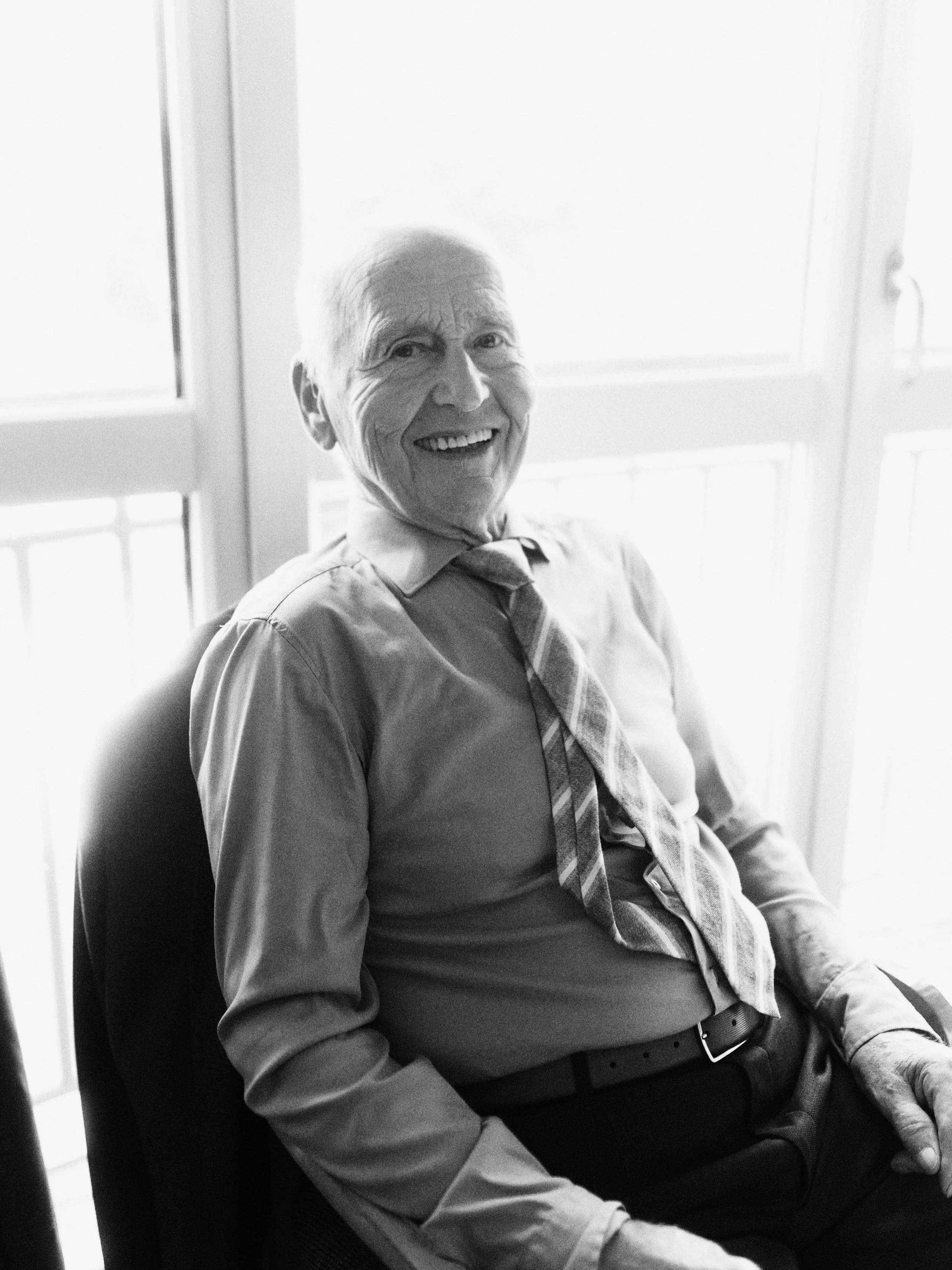 Schwarz-Weiß-Foto eines lächelnden älteren Mannes, der in einem Hemd und Krawatte sitzt, vor einem Fenster.