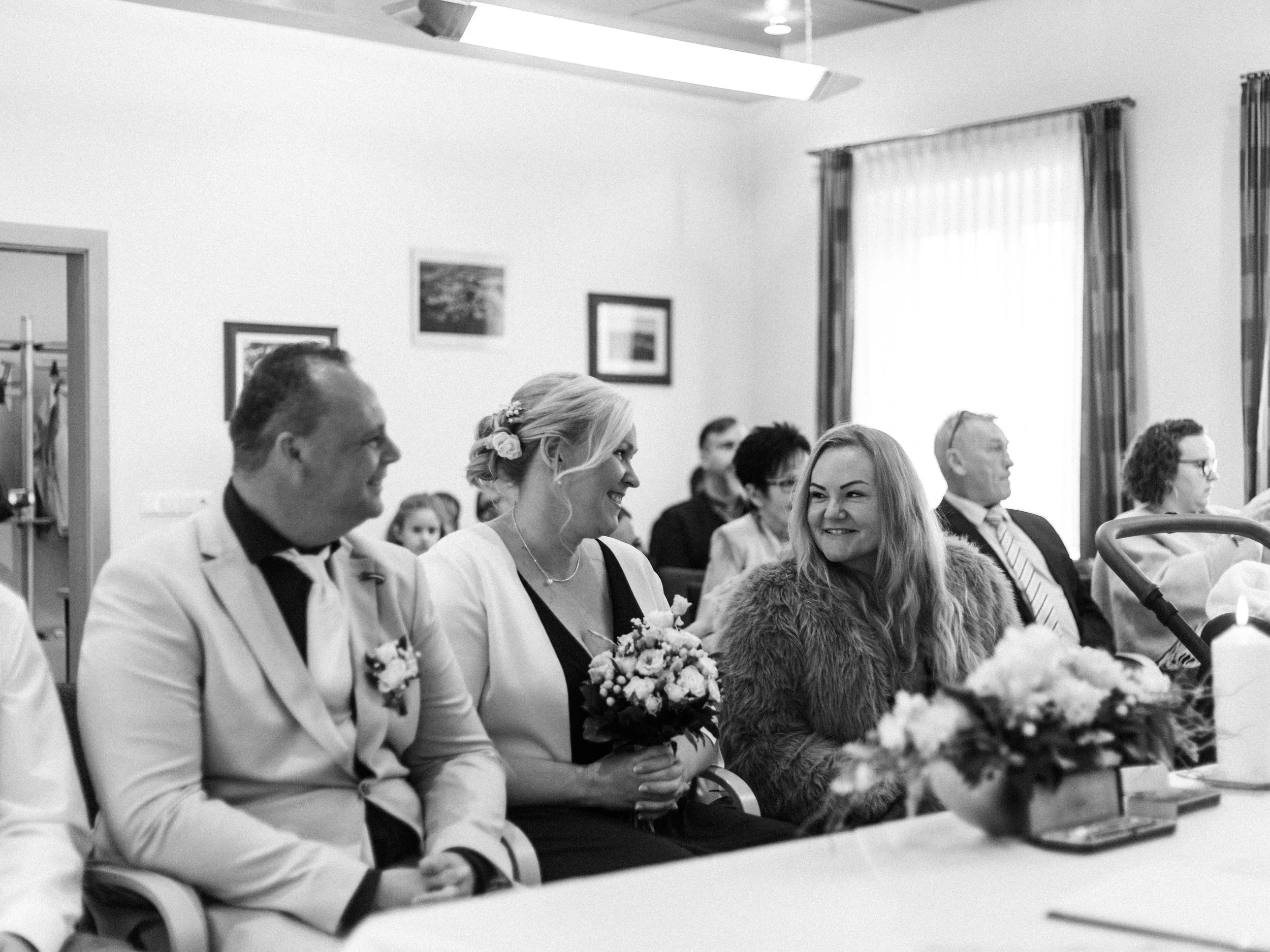 Hochzeitspaar sitzt in einem Saal, umgeben von Gästen, bei der Zeremonie. Die Braut hält einen Blumenstrauß, alle lachen und genießen den Moment.