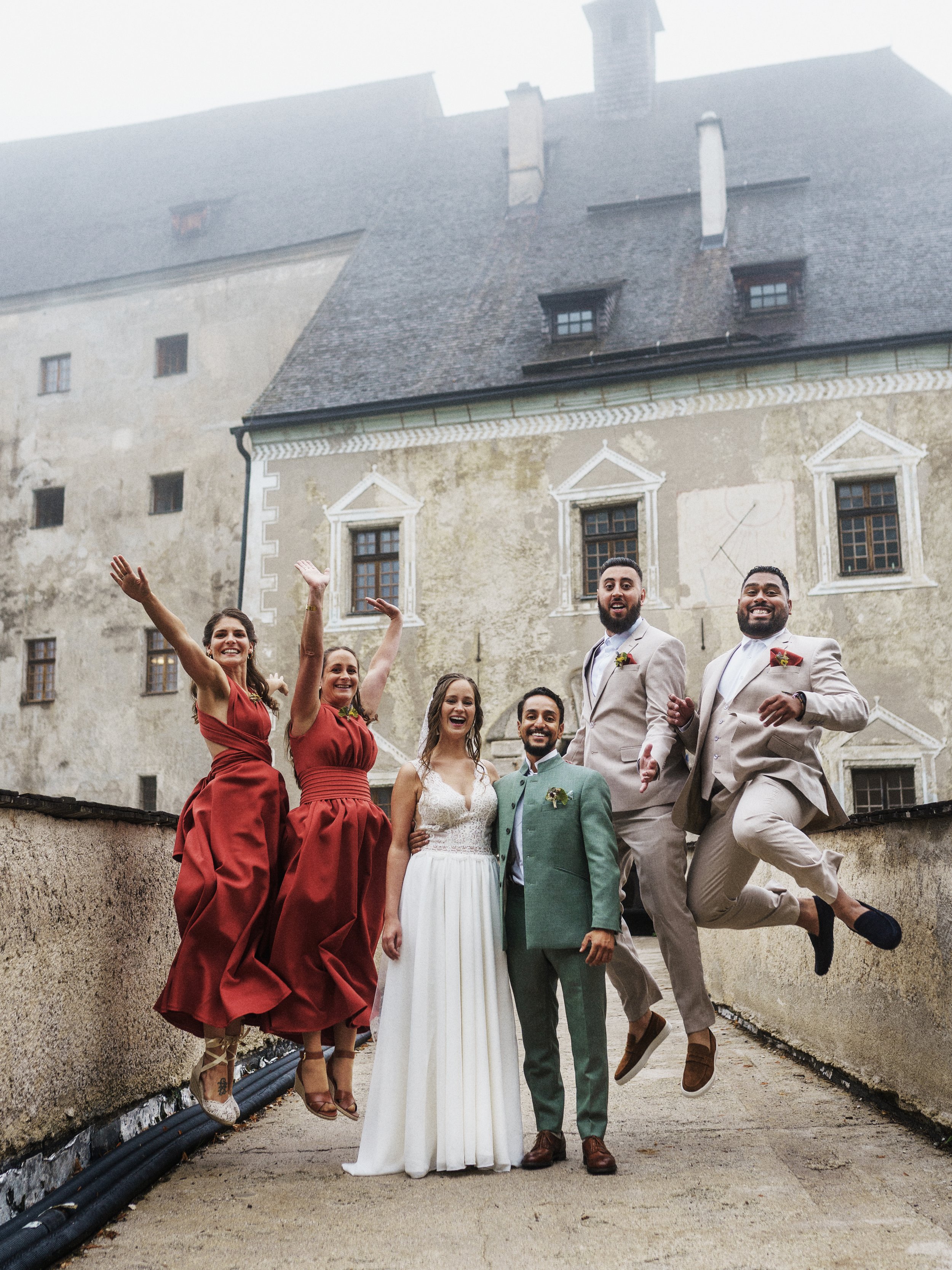 Hochzeitspaar und Freunde posieren vor einem alten Gebäude, einige springen, alle lachen und haben Spaß.