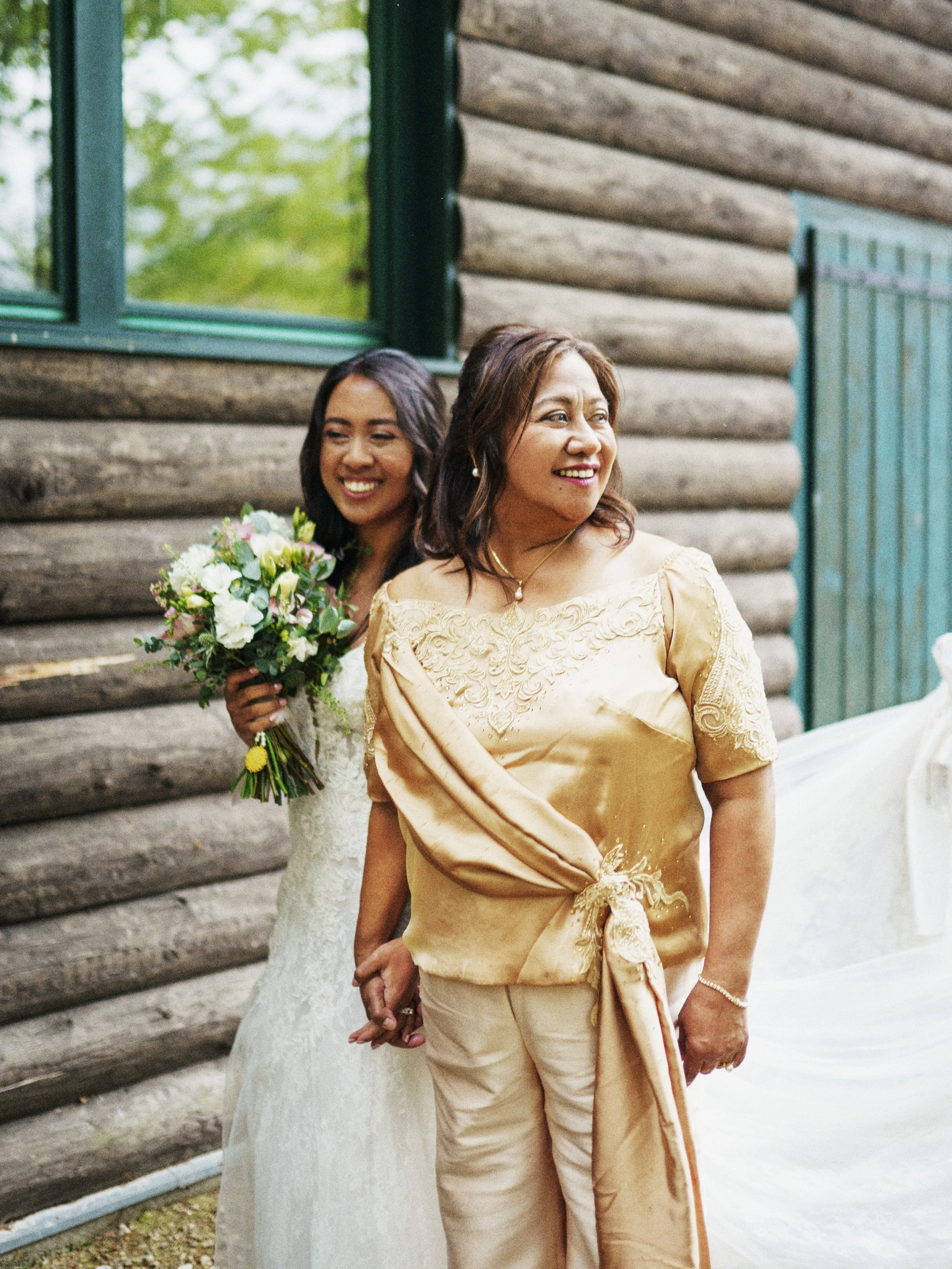 Zwei Frauen, eine in einem weißen Brautkleid mit Blumenstrauß, die andere in einem goldfarbenen Kleid, stehen vor einem Holzhaus und halten Händchen, während sie nach vorne schauen.