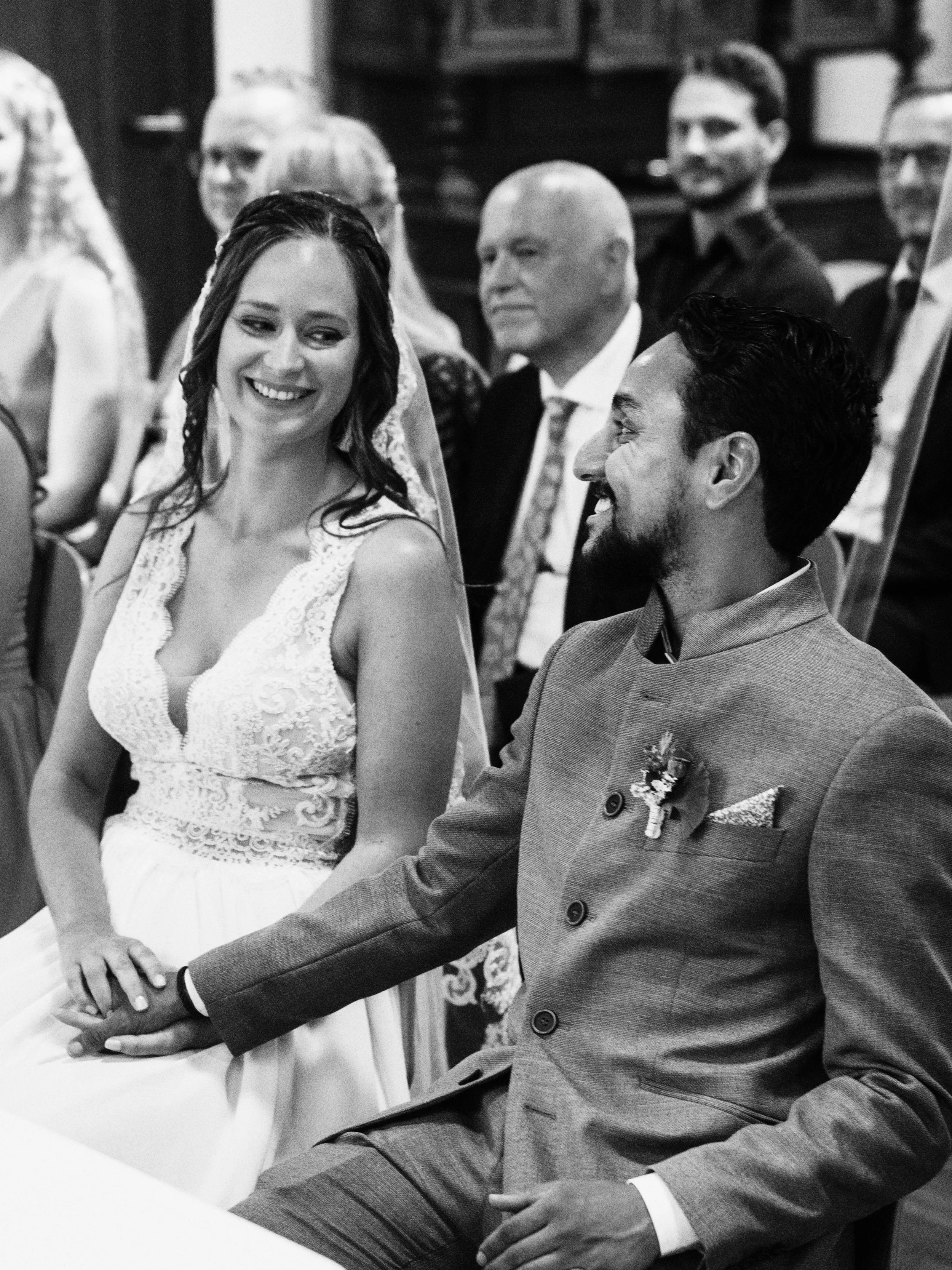 Braut und Bräutigam sitzen bei einer Hochzeitszeremonie, der Bräutigam hält die Hand der Braut. Sie lachen sich an, während die Gäste im Hintergrund zuschauen.