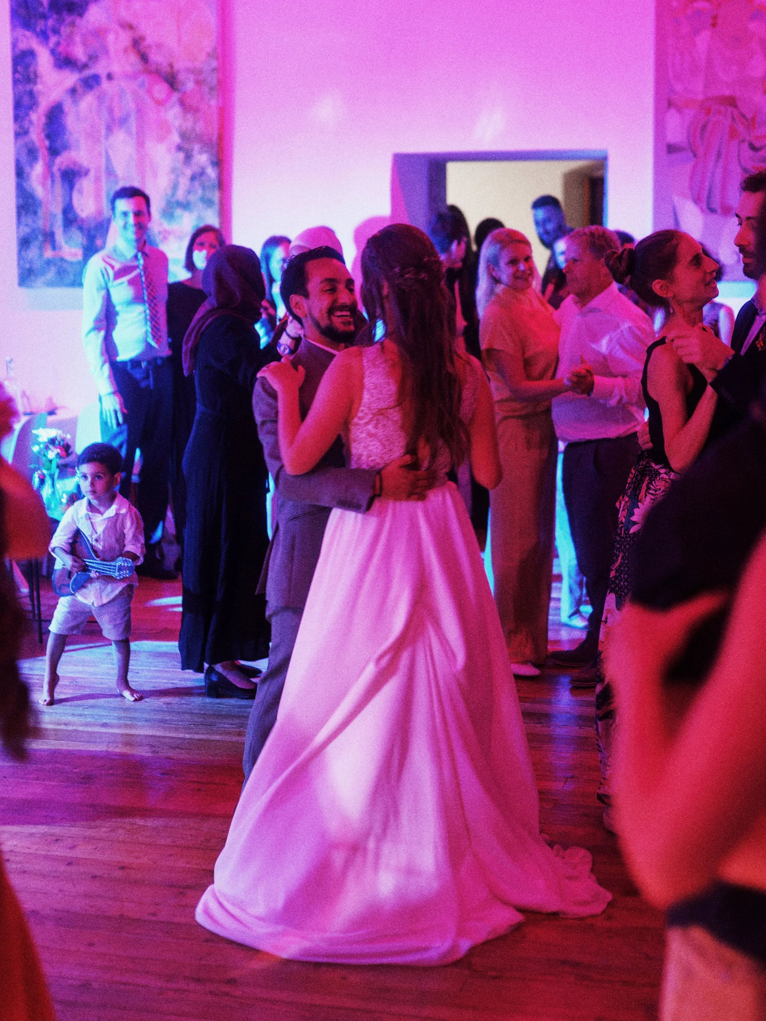 Braut und Bräutigam tanzen auf einer festlichen Hochzeit, umgeben von Gästen, im Hintergrund sind bunte Bilder an der Wand und ein Kind, das ein Gitarren-Spiel spielt.