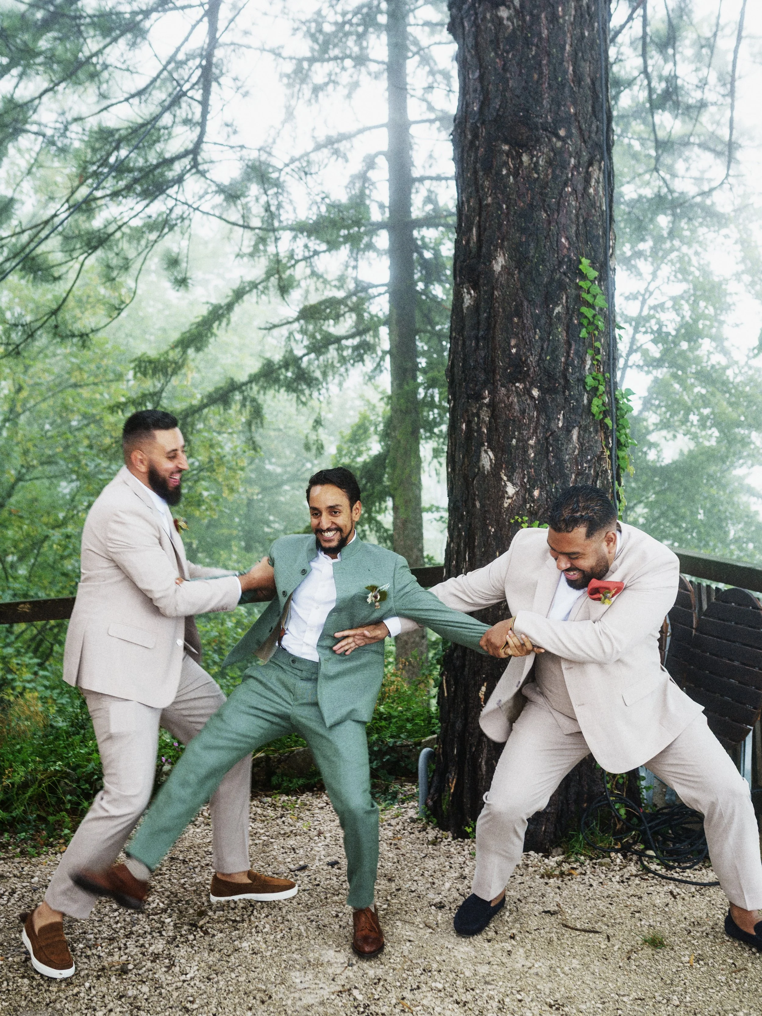 Drei Männer in formeller Kleidung spielen im Wald, lachen und ziehen sich an den Armen. Es ist ein grüner, bewaldeter Hintergrund mit hohen Bäumen.