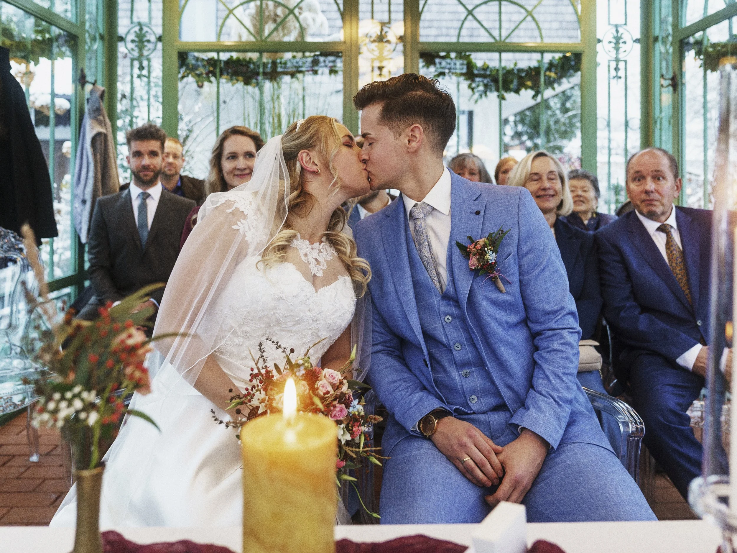 Ein Brautpaar küsst sich während ihrer Hochzeitszeremonie, umgeben von Freunden und Familie in einem hellen, grünen Gewächshaus.