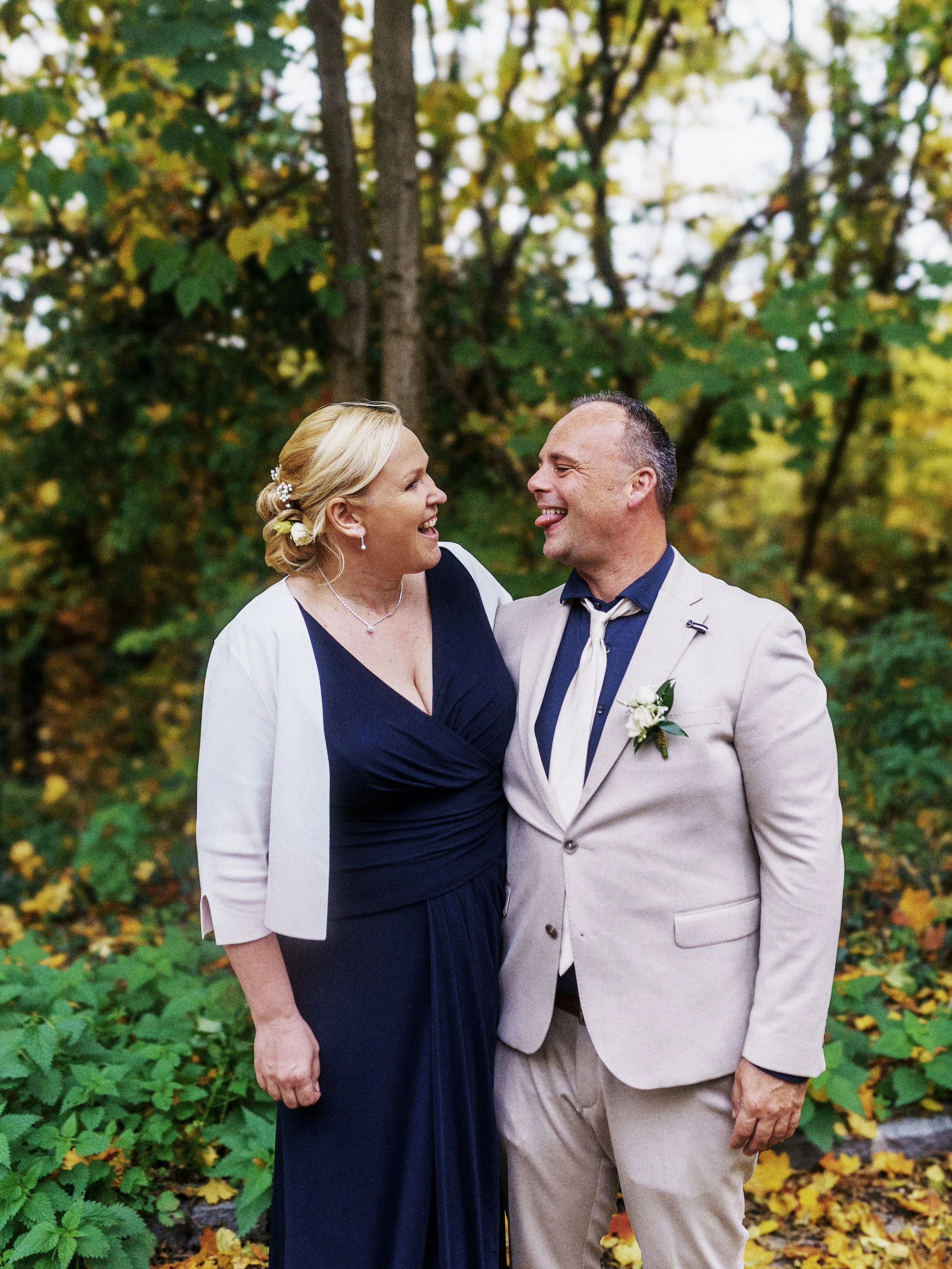 Ein lachendes Paar beim Außentermin in einem Wald, vermutlich bei einer Hochzeit, umgeben von herbstlicher Laubfärbung.