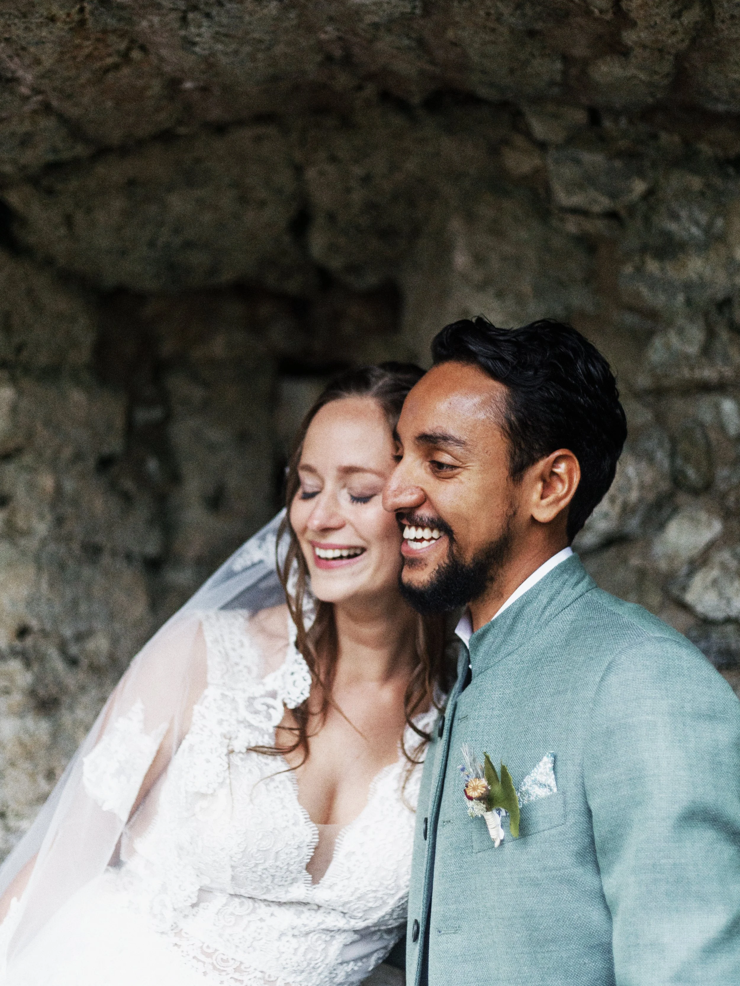 Ein lachendes Brautpaar mit einem Mann in einem grünen Anzug und einer Frau in einem weißen Hochzeitskleid vor einer steinernen Wand.