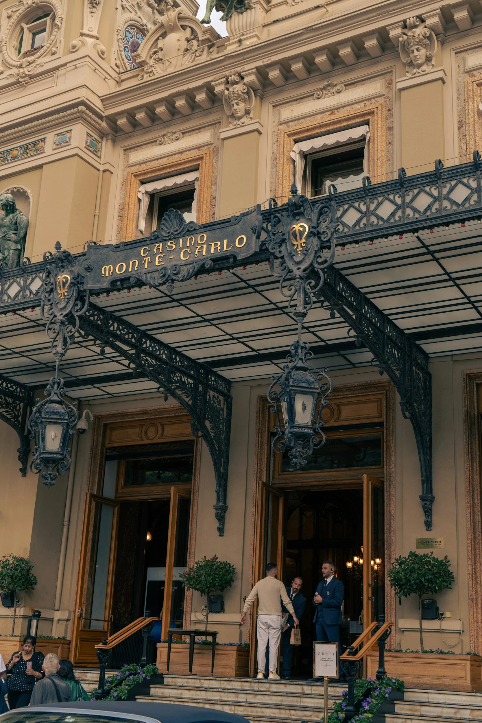 Exterior of a building with ornate architectural details, including sculptures and decorative ironwork, bearing a sign that reads 'Casino Monte Carlo'. The entrance has open wooden doors, with people standing and talking outside.