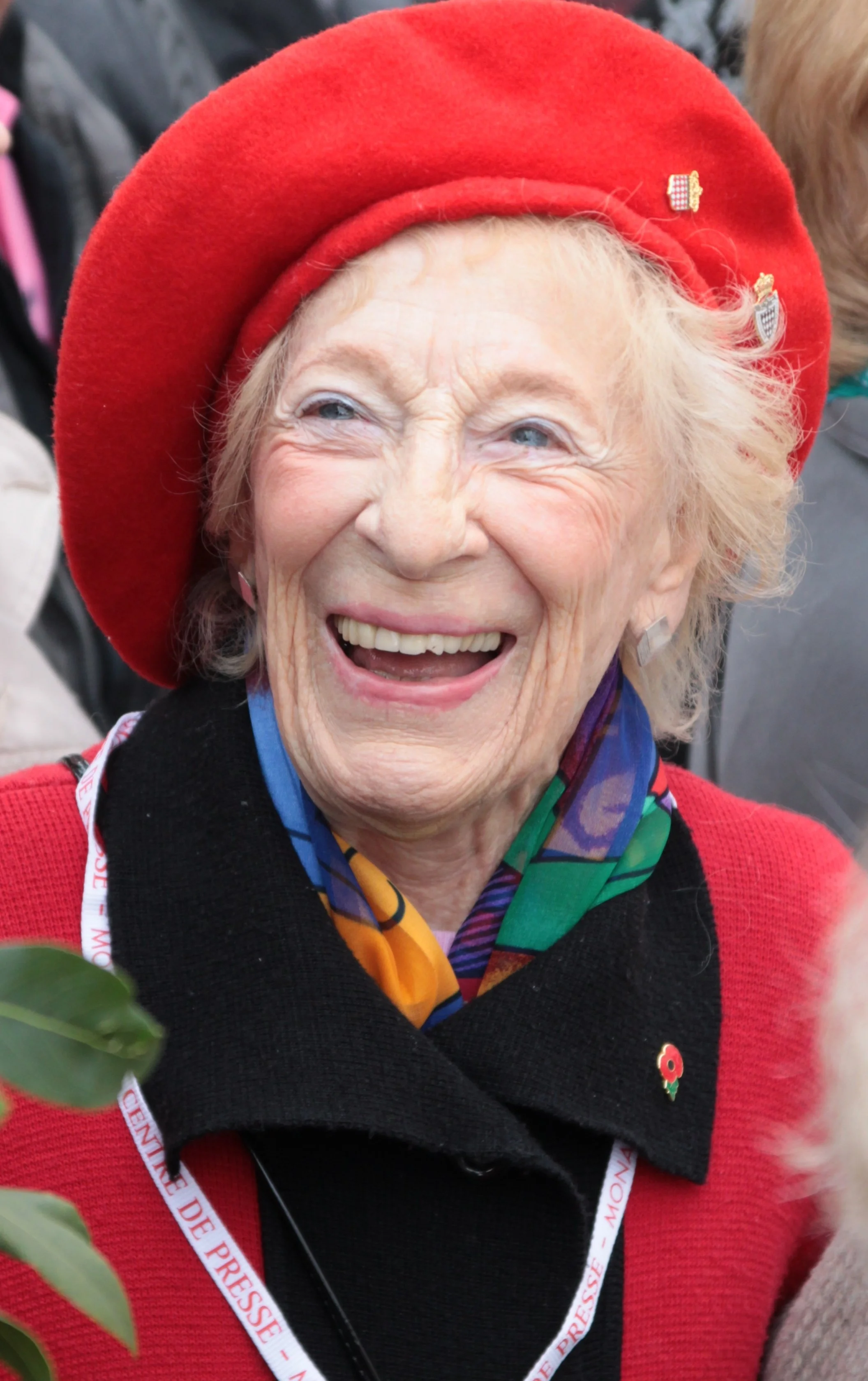 Molly Brown, woman with a big smile, wearing a red hat and a black and red coat, colorful scarf, and a poppy pin, at an outdoor event.