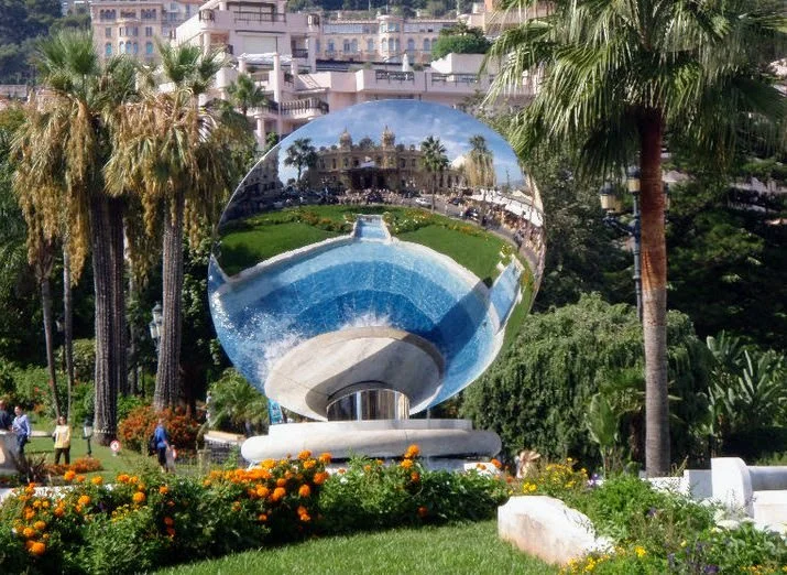 A large reflective spherical sculpture in a park with palm trees and flowers, reflecting a historic building and people walking.