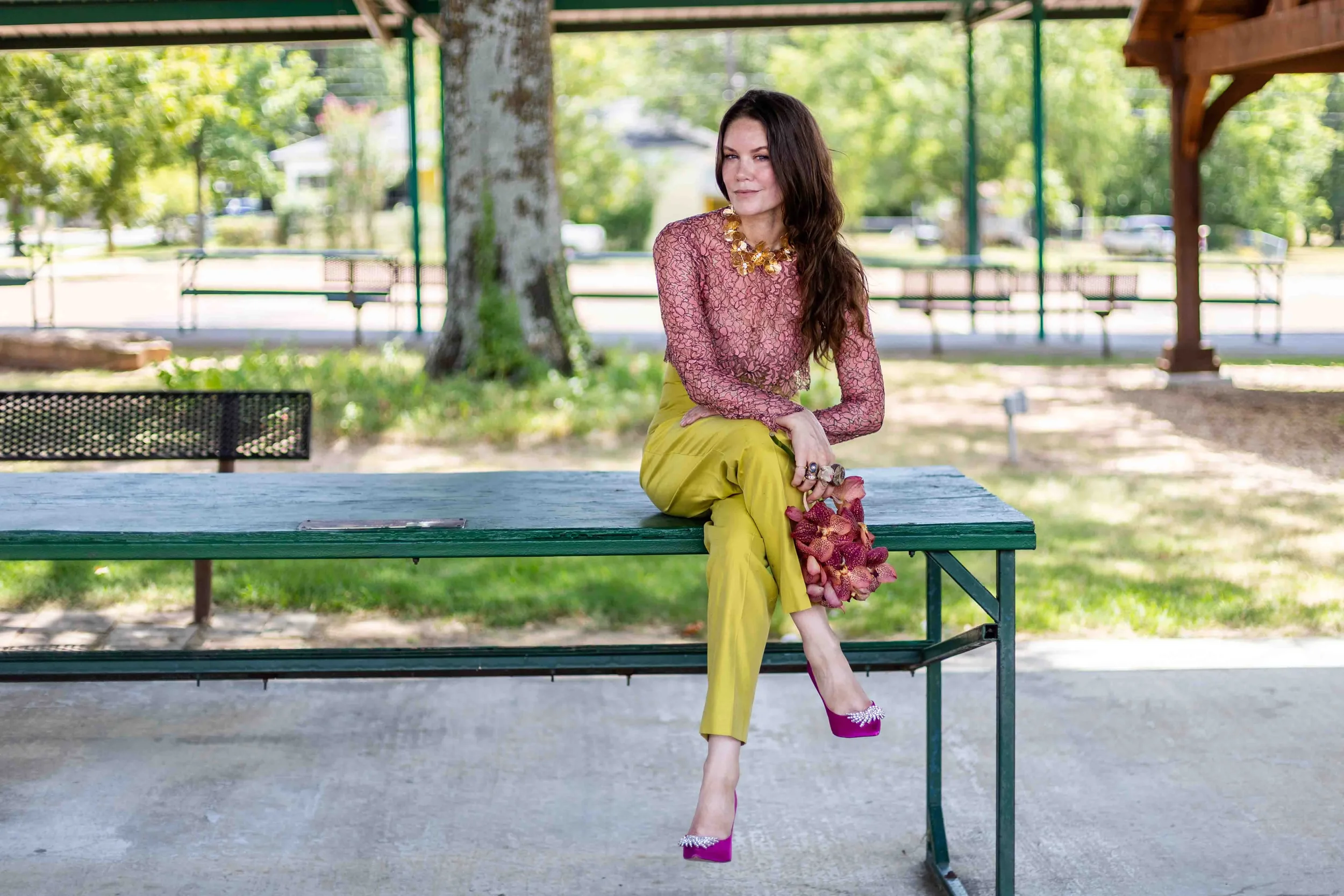 A woman sitting on a park bench under a shelter, wearing a pink lace top, yellow pants, and purple high-heeled shoes with rhinestones, holding a bunch of pink orchids, with green trees and benches in the background.
