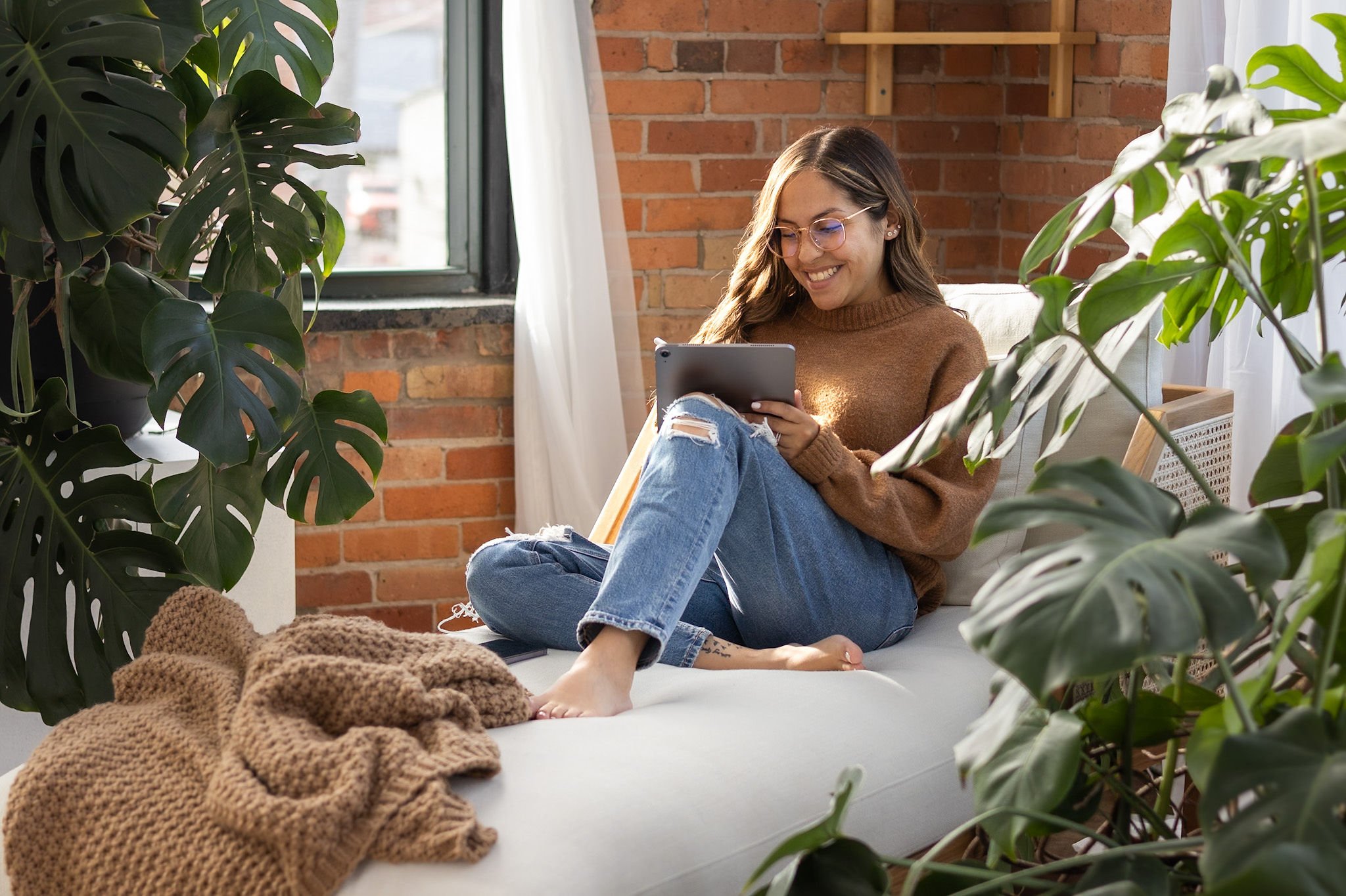 A young woman with glasses and long brown hair sits comfortably on a white sofa, smiling as she looks at a tablet. She wears a brown sweater and ripped jeans. The room has a brick wall and large windows with sheer white curtains. Green houseplants surround her, creating a cozy and vibrant atmosphere.