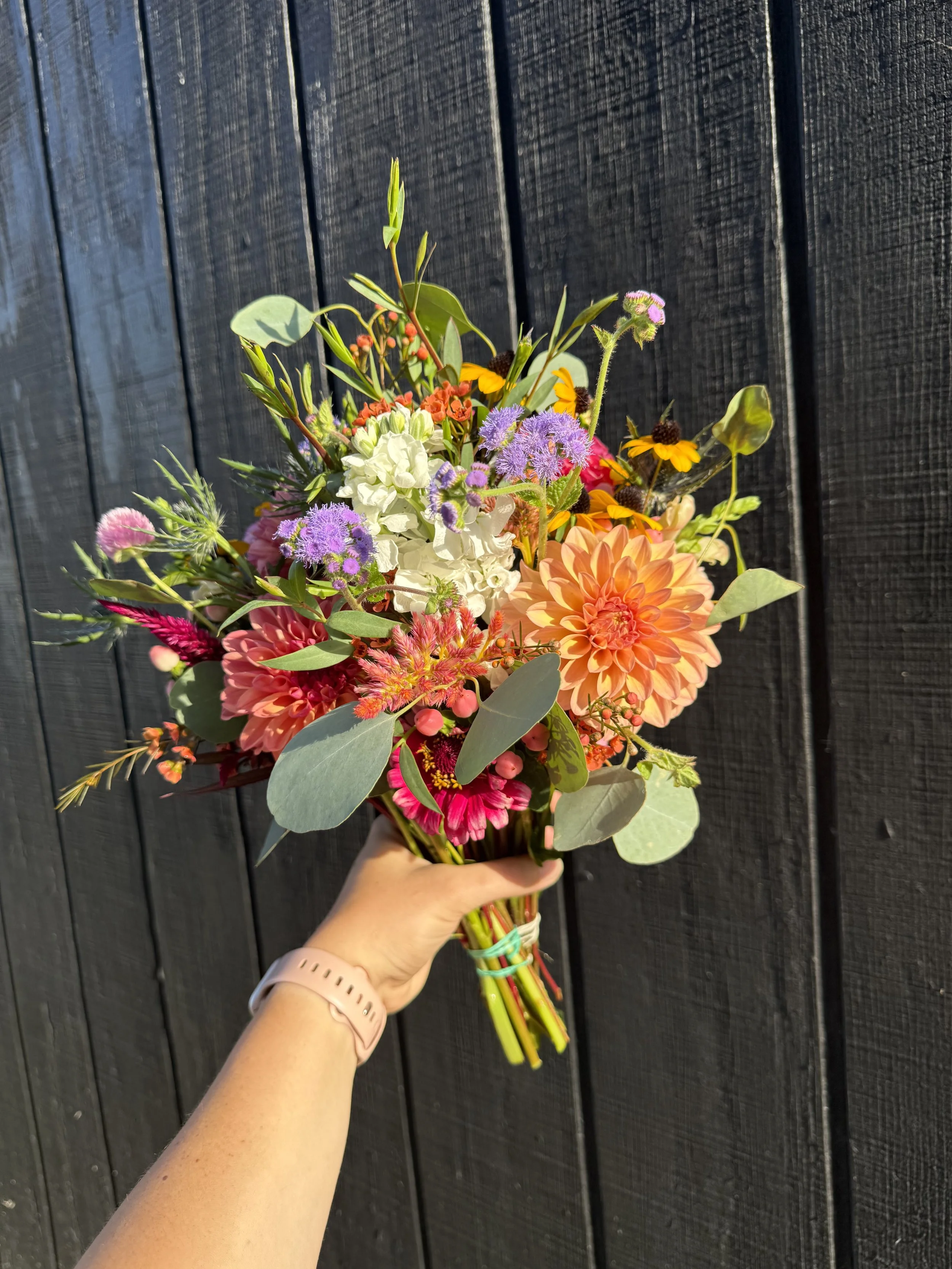 A colorful bouquet of flowers held by a person's hand in front of a black wooden wall.