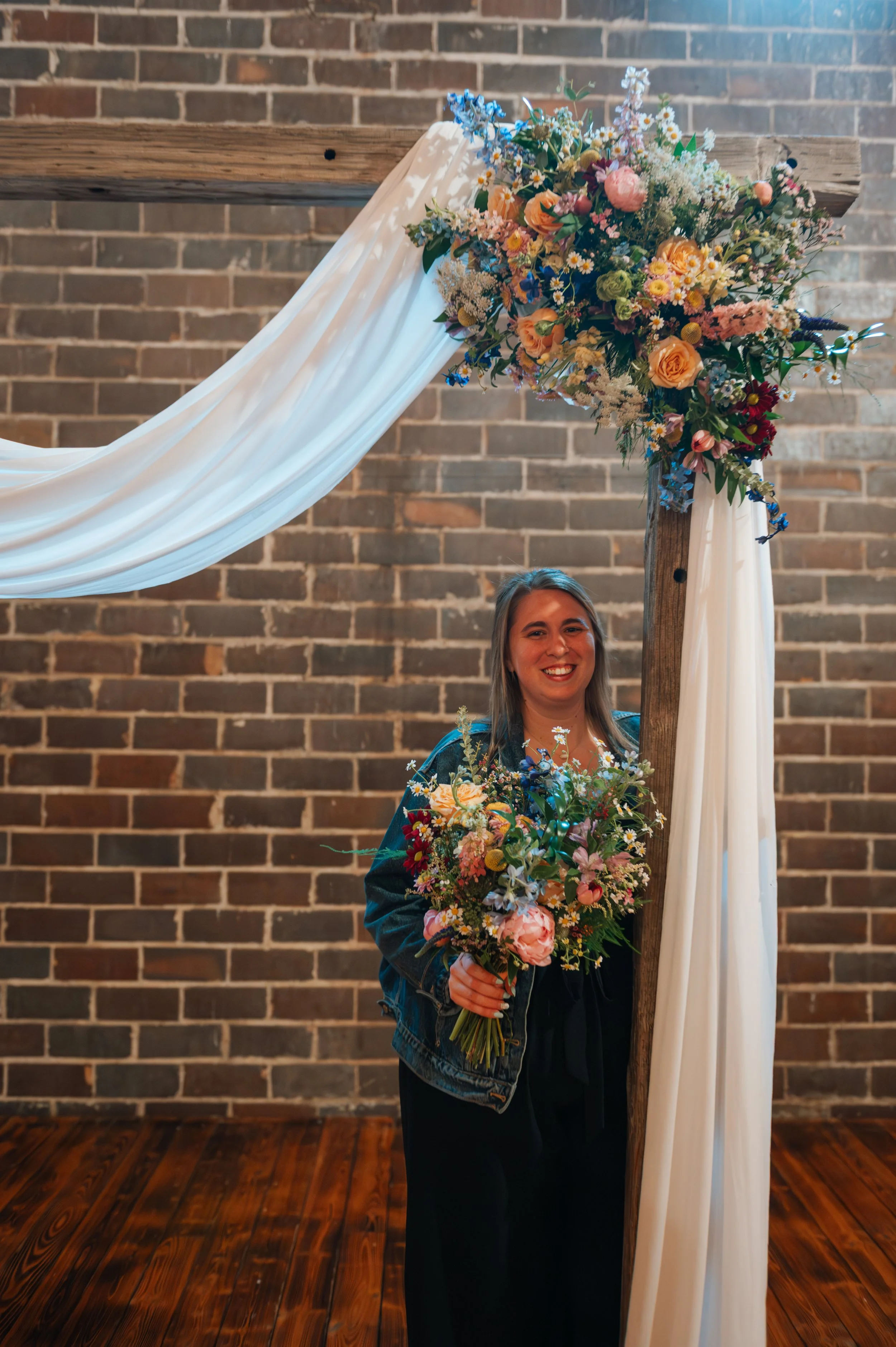 A woman holding a bouquet of flowers, standing under a floral arch decorated with flowing white fabric and various colorful flowers, against a brick wall background.