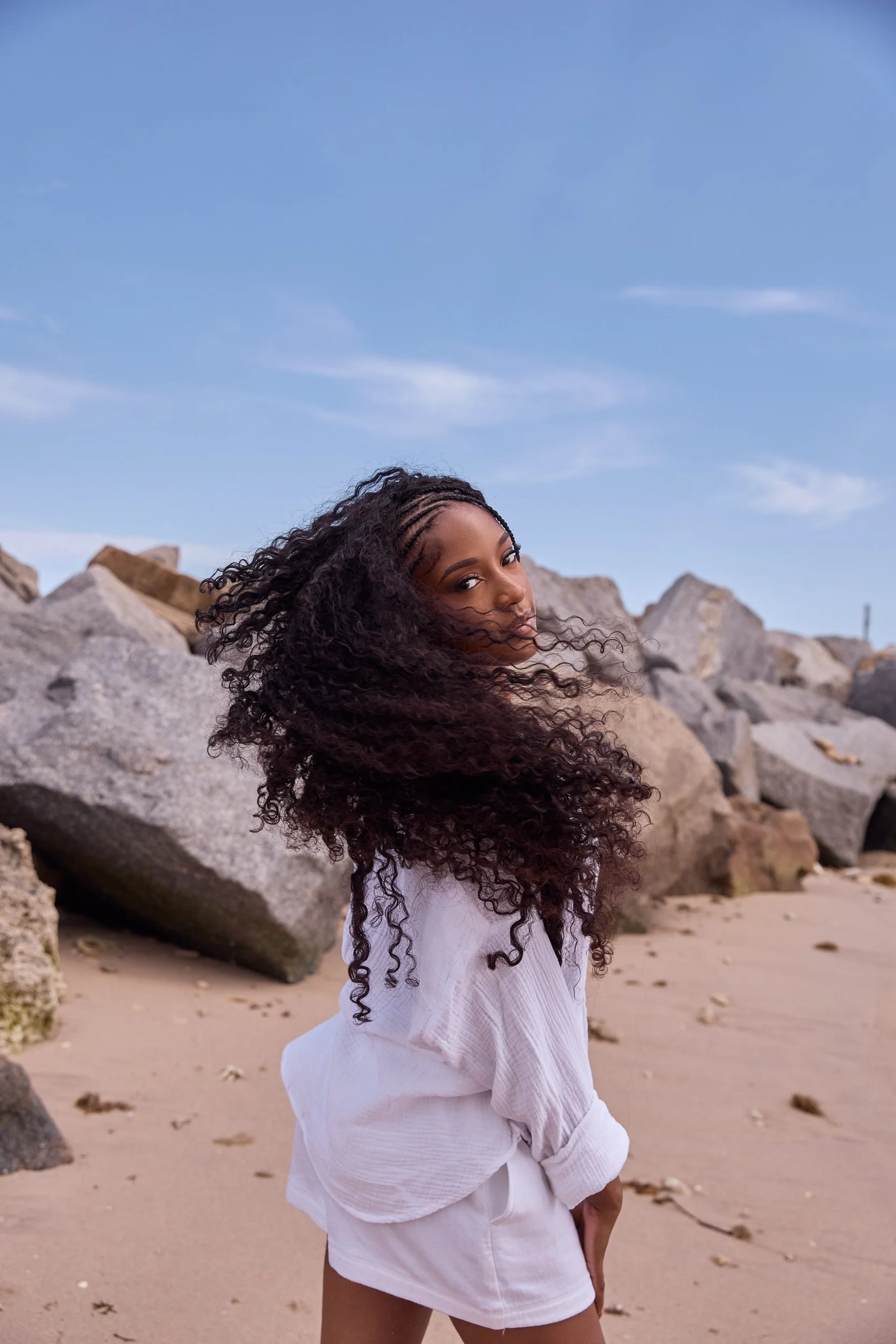 A woman with curly hair standing on a beach with large rocks behind her, wearing a white shirt and white shorts, looking at the camera with a blue sky overhead.