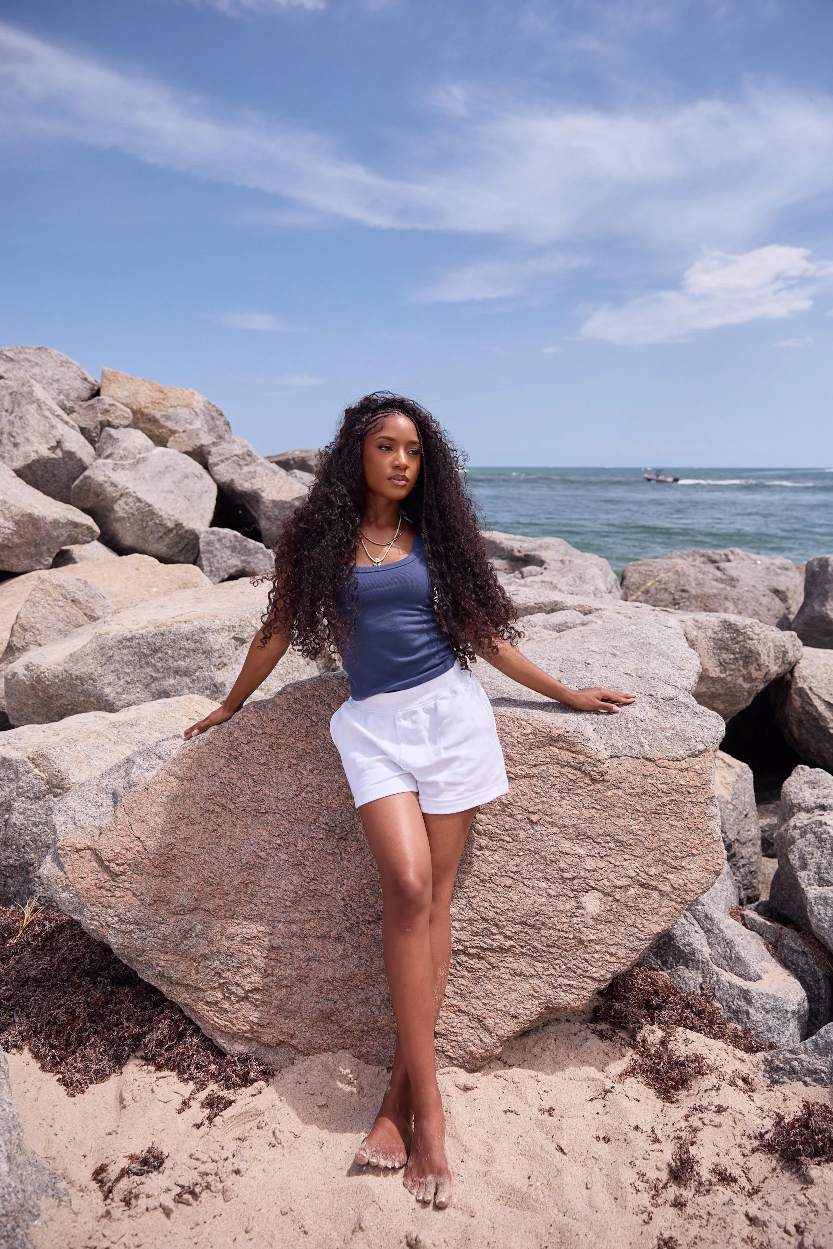 A woman in a blue shirt and white shorts standing on rocks at the beach with the ocean and a boat in the background.