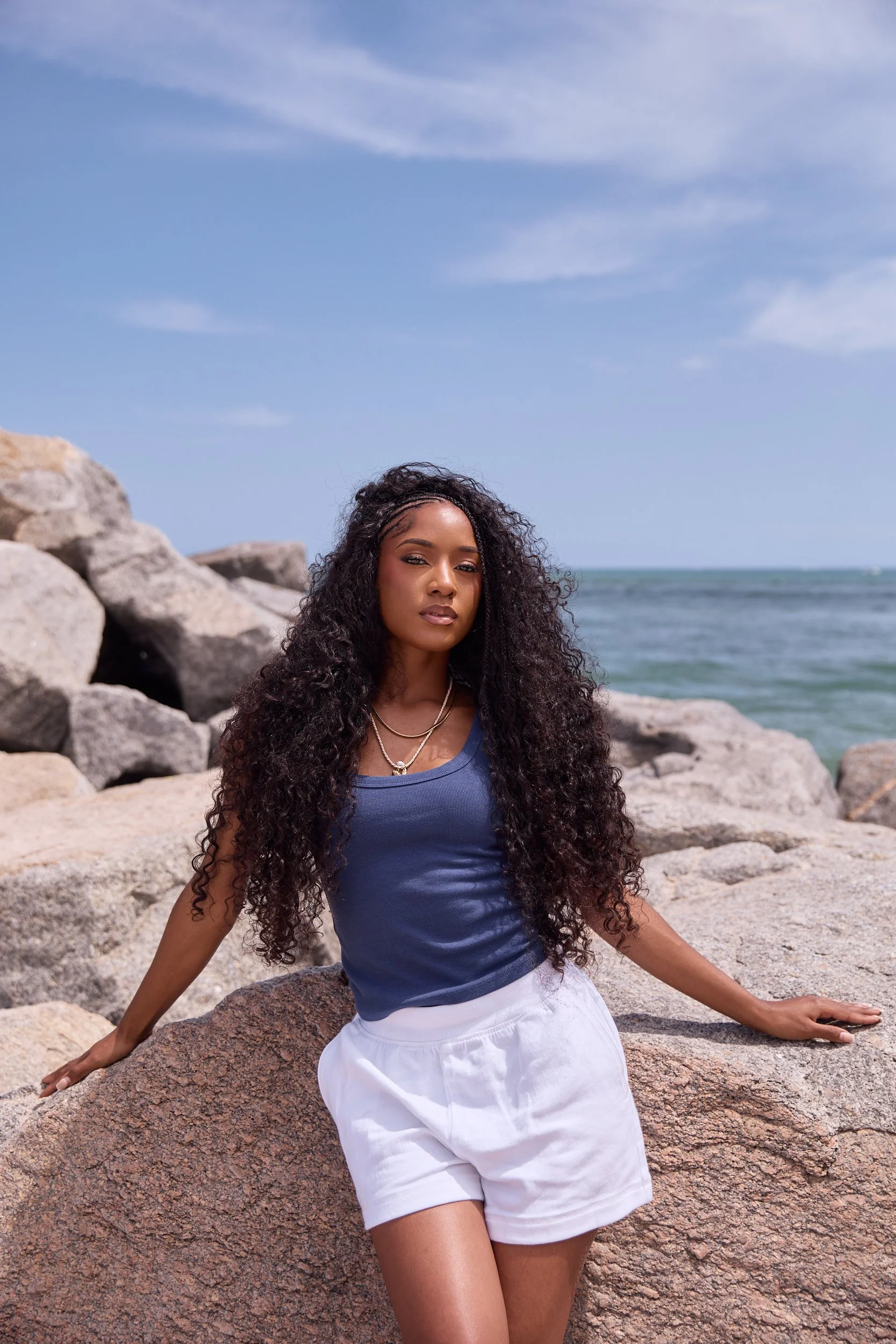 A woman with long curly hair and jewelry in a blue top and white shorts sitting on a rocky beach with large rocks, the ocean, and a partly cloudy sky in the background.