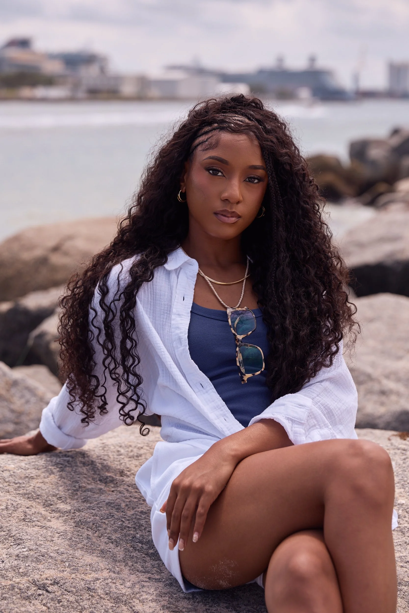 A woman with long, curly black hair and gold hoop earrings sitting on a rocky beach, wearing a navy blue top and white shirt, with sunglasses hanging from her shirt collar, is looking at the camera with a serious expression, with water and boats in t