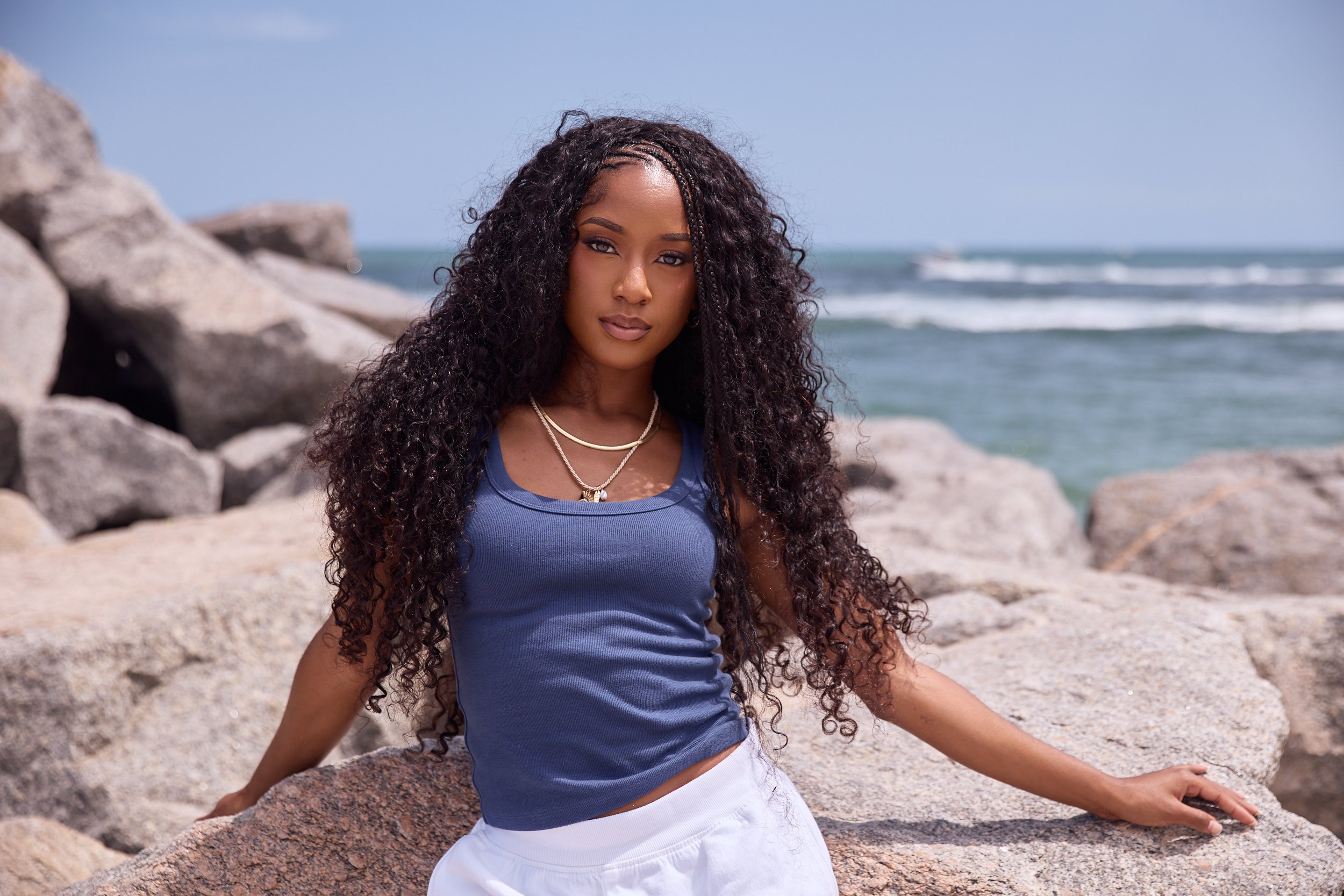 Woman with long curly hair sitting on rocks at the beach, wearing a blue tank top and white skirt, with ocean waves in the background.